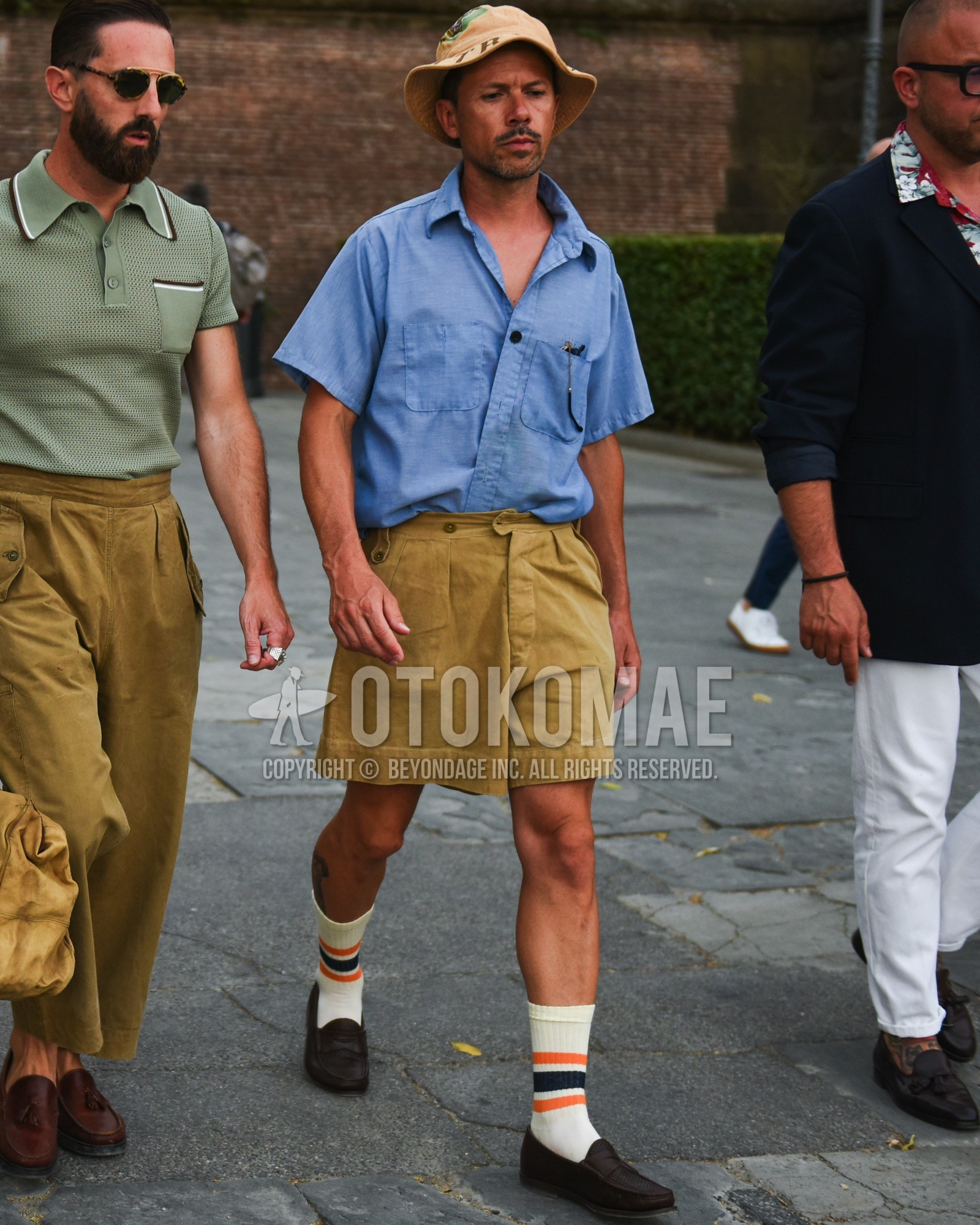 Men's spring summer outfit with beige graphic bucket hat, blue plain shirt, beige plain short pants, white horizontal stripes socks, brown coin loafers leather shoes.