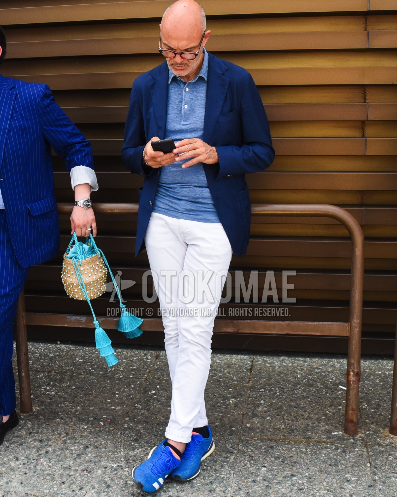 Men's spring summer outfit with brown tortoiseshell glasses, navy plain tailored jacket, blue plain polo shirt, white plain cotton pants, black plain socks, blue low-cut sneakers.