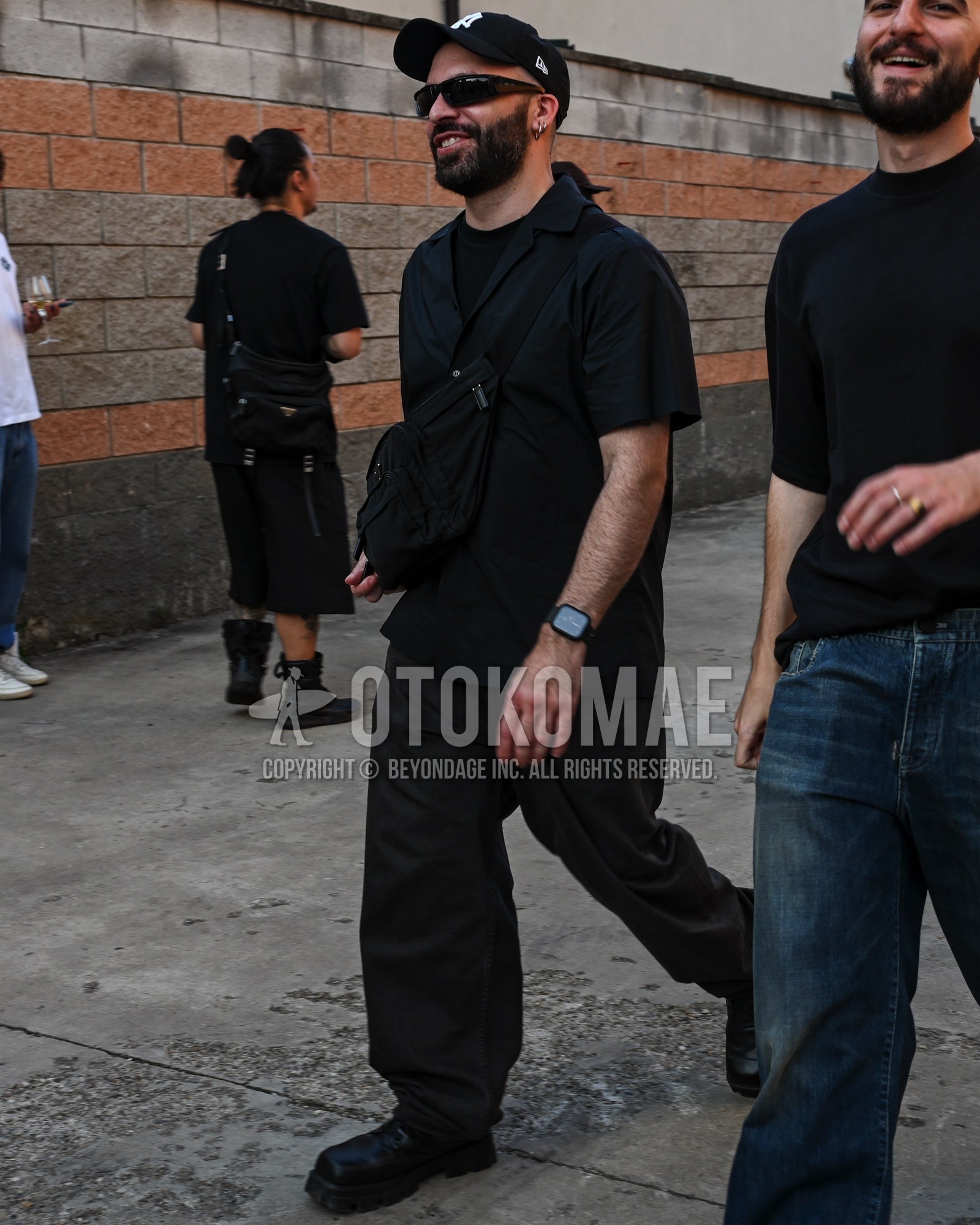 Men's spring summer outfit with black deca logo baseball cap, black plain t-shirt, black plain shirt, black plain wide pants, black work boots, black plain shoulder bag.