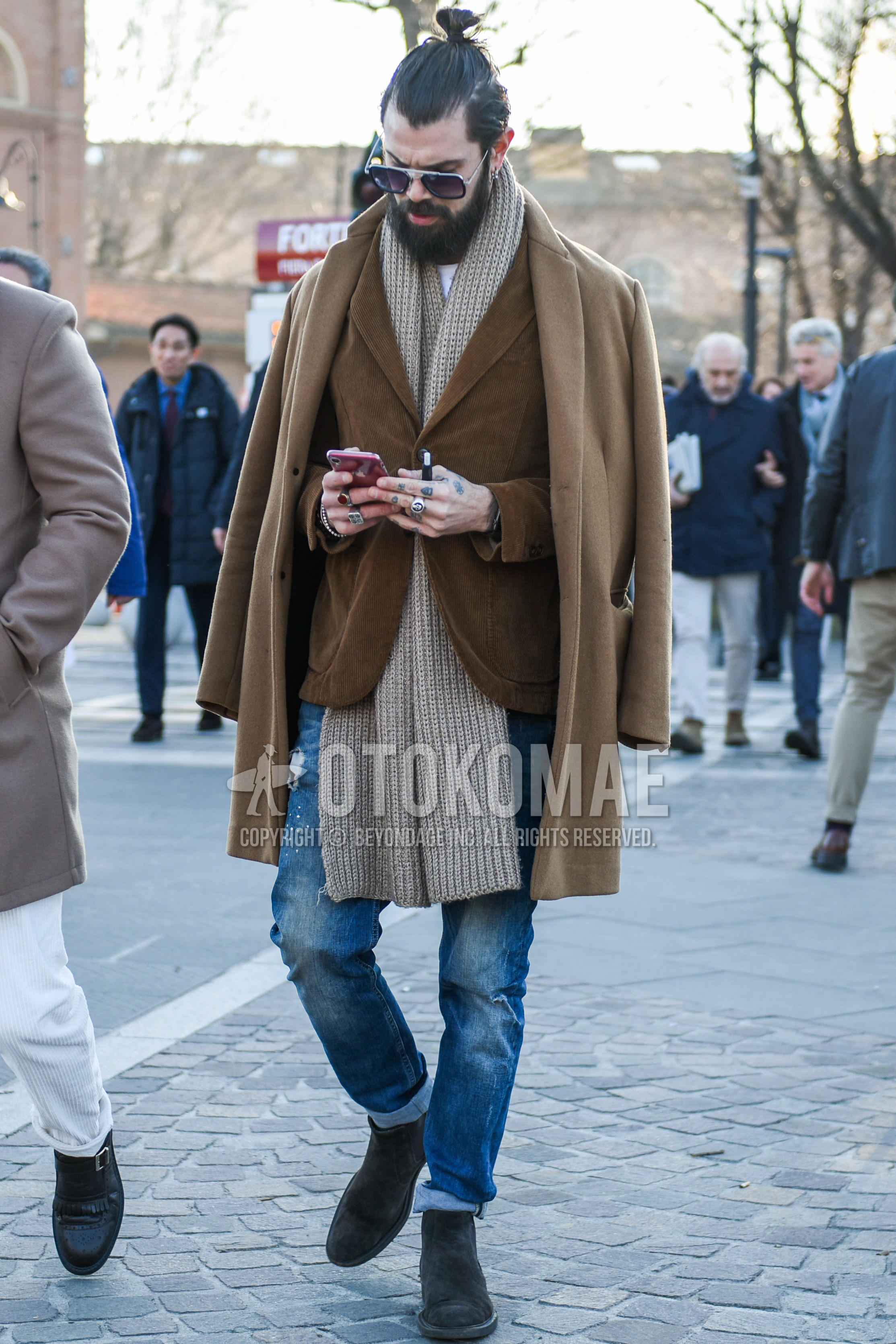 Men's autumn winter outfit with black silver plain glasses, beige plain scarf, beige plain chester coat, brown plain tailored jacket, blue plain damaged jeans, gray side-gore boots.