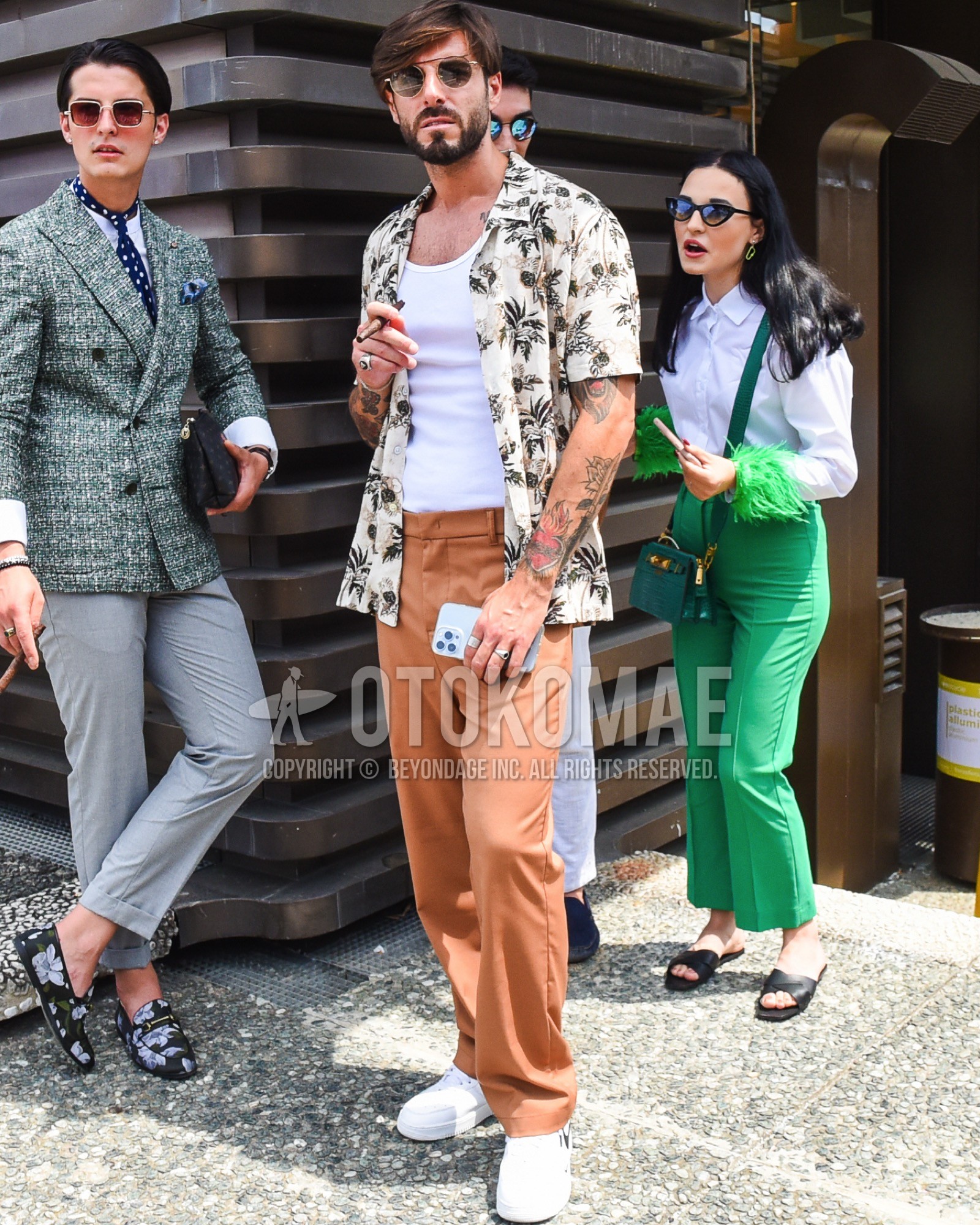 Men's spring summer outfit with gold plain sunglasses, white plain tank top, beige botanical shirt, brown plain slacks, white low-cut sneakers.