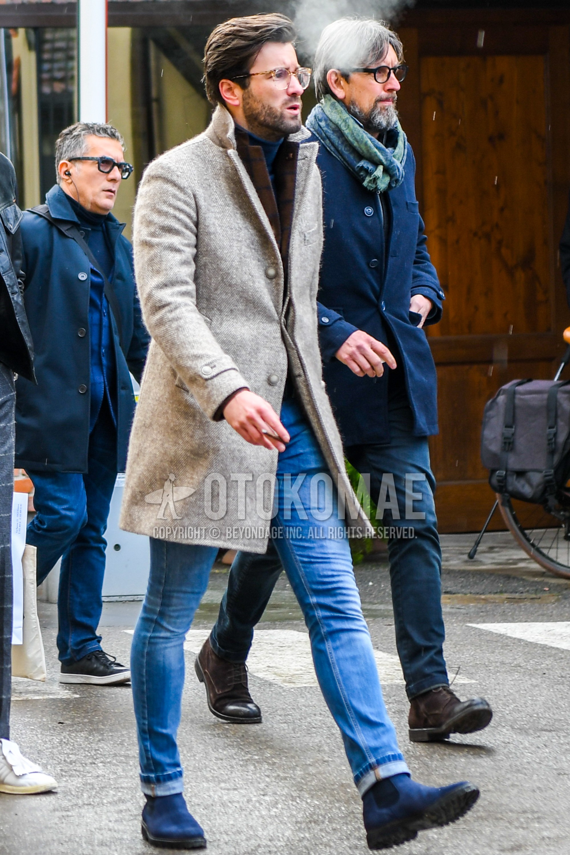 Men's autumn winter outfit with plain glasses, beige plain chester coat, brown plain tailored jacket, gray plain turtleneck knit, blue plain denim/jeans, blue side-gore boots.
