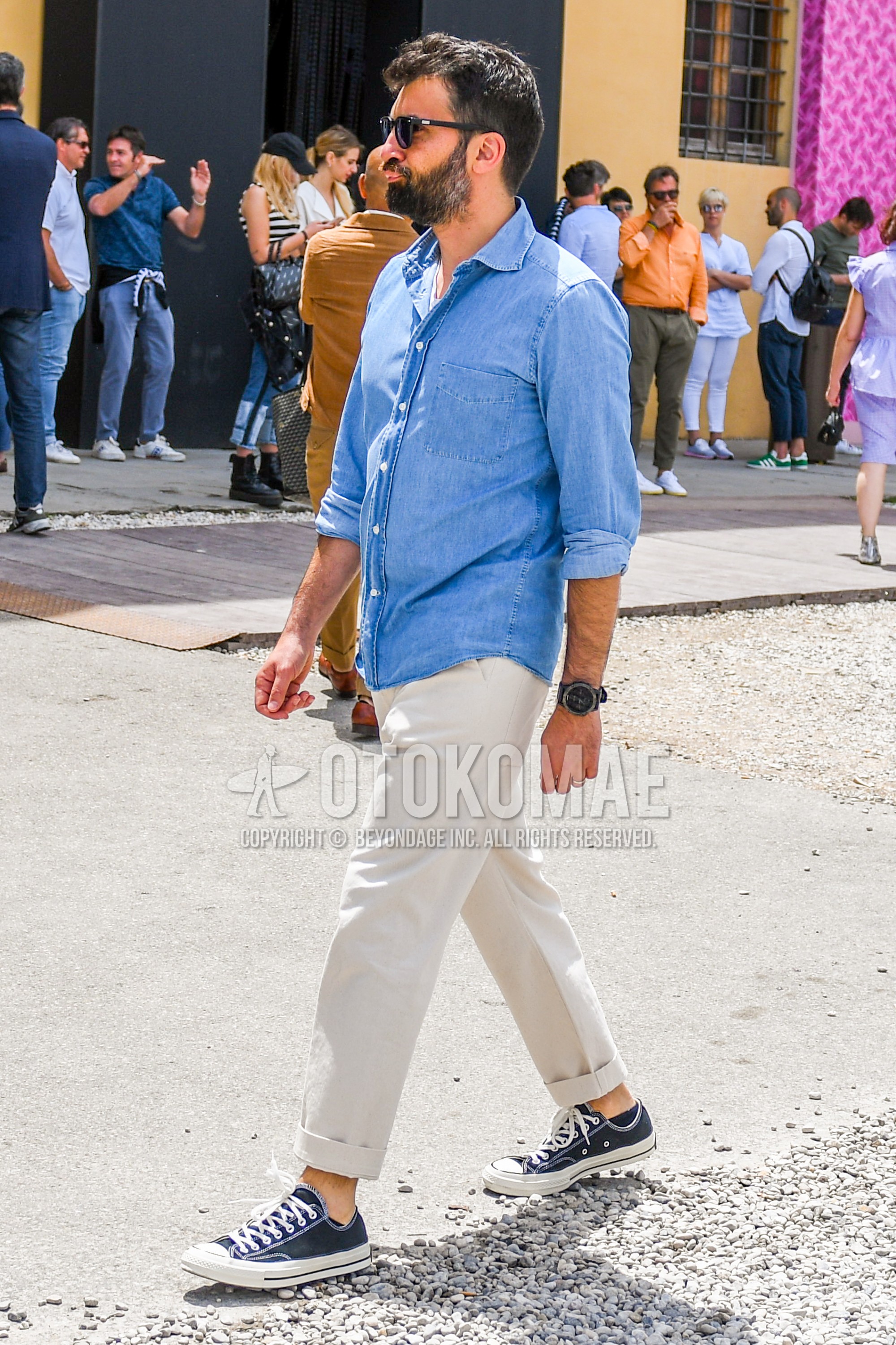 Men's spring summer outfit with plain sunglasses, blue plain denim shirt/chambray shirt, beige plain chinos, navy low-cut sneakers.