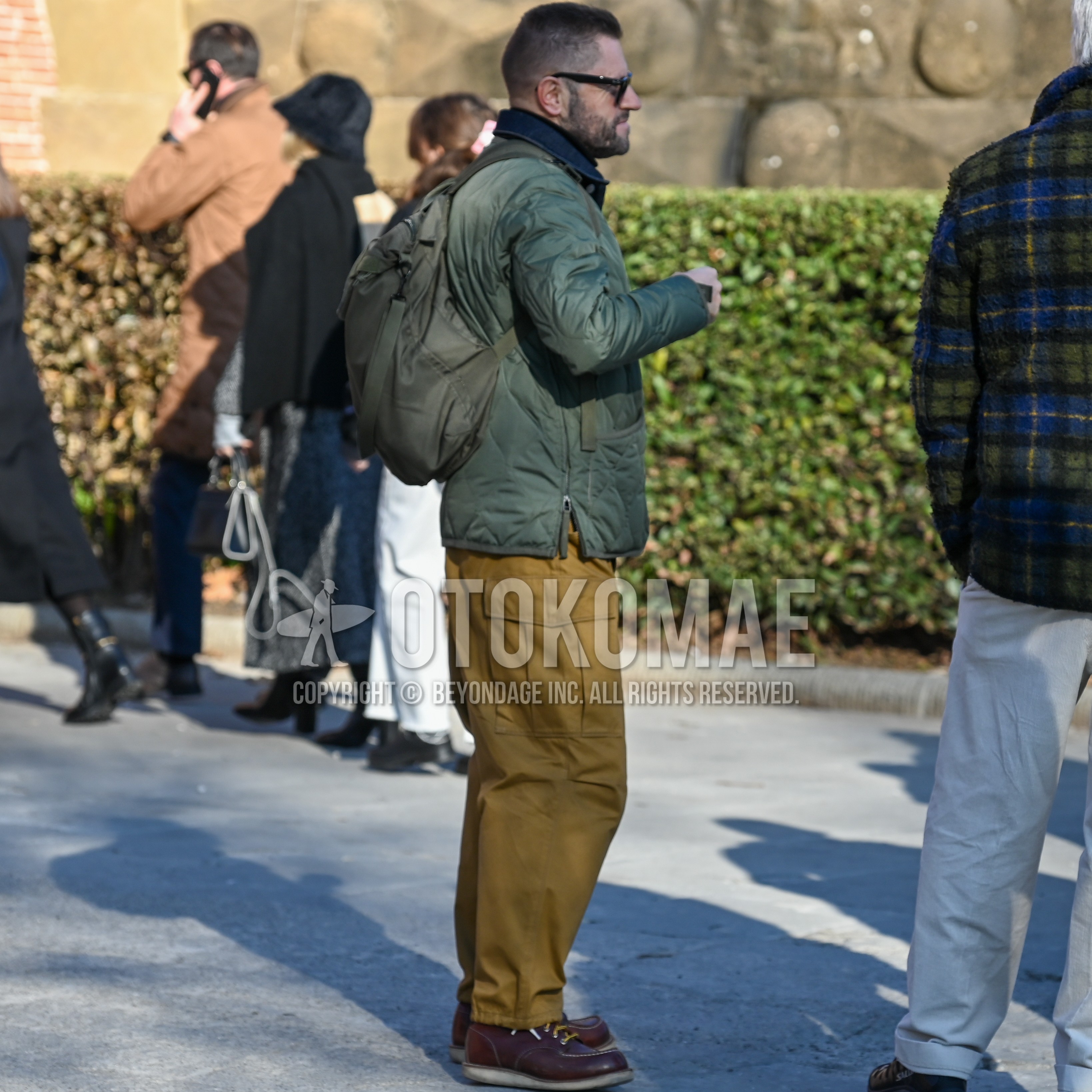 Men's autumn winter outfit with black plain sunglasses, olive green plain quilted jacket, brown plain work pants, brown work boots, olive green plain backpack.