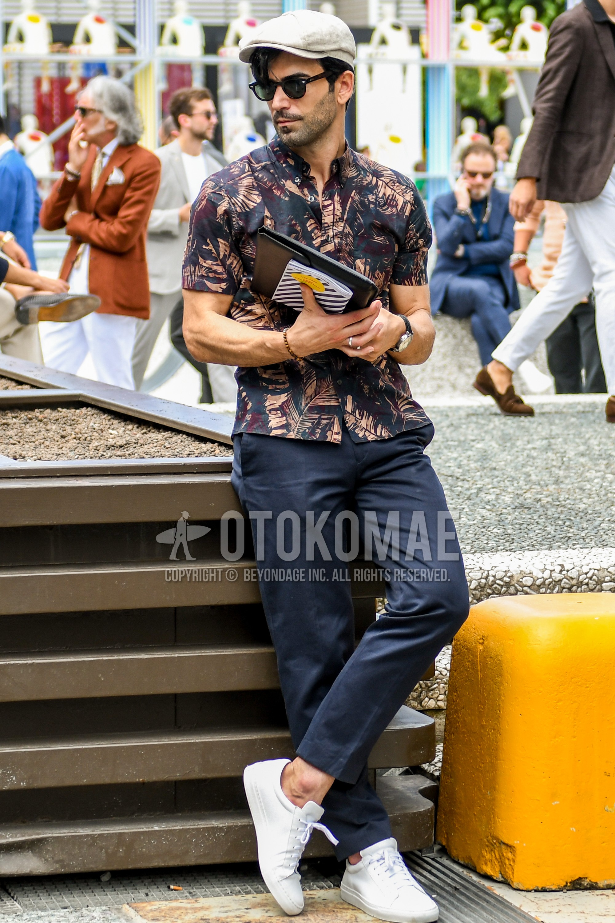 Men's summer outfit with beige plain cap, plain sunglasses, multi-color botanical shirt, navy plain ankle pants, white low-cut sneakers.