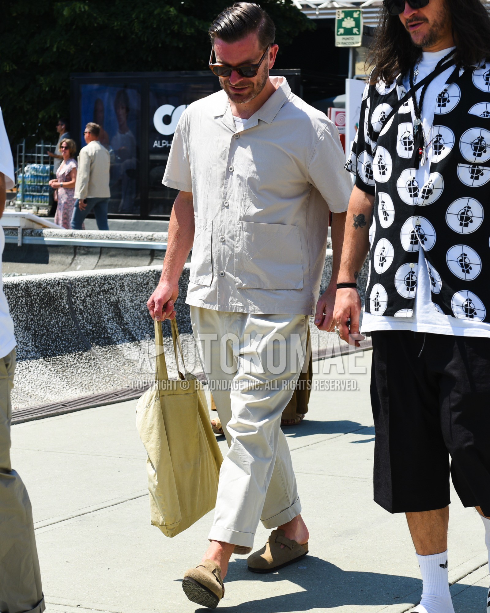Men's spring summer outfit with brown tortoiseshell sunglasses, beige plain shirt, white plain t-shirt, beige plain cotton pants, beige plain cropped pants, beige leather sandals, beige plain tote bag.