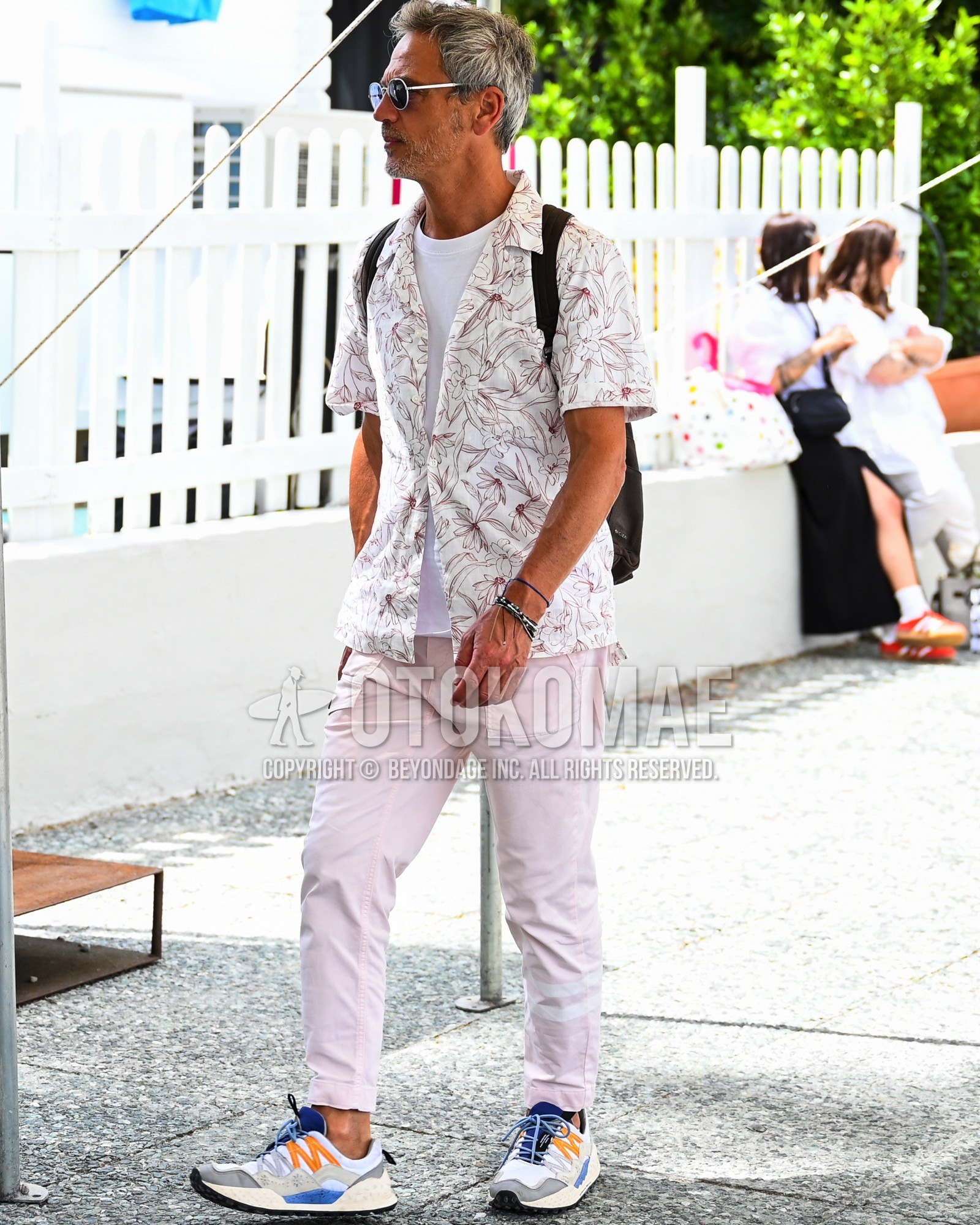 Men's spring summer outfit with black plain sunglasses, white whole pattern shirt, white plain t-shirt, pink plain chinos, white low-cut sneakers, black plain backpack.