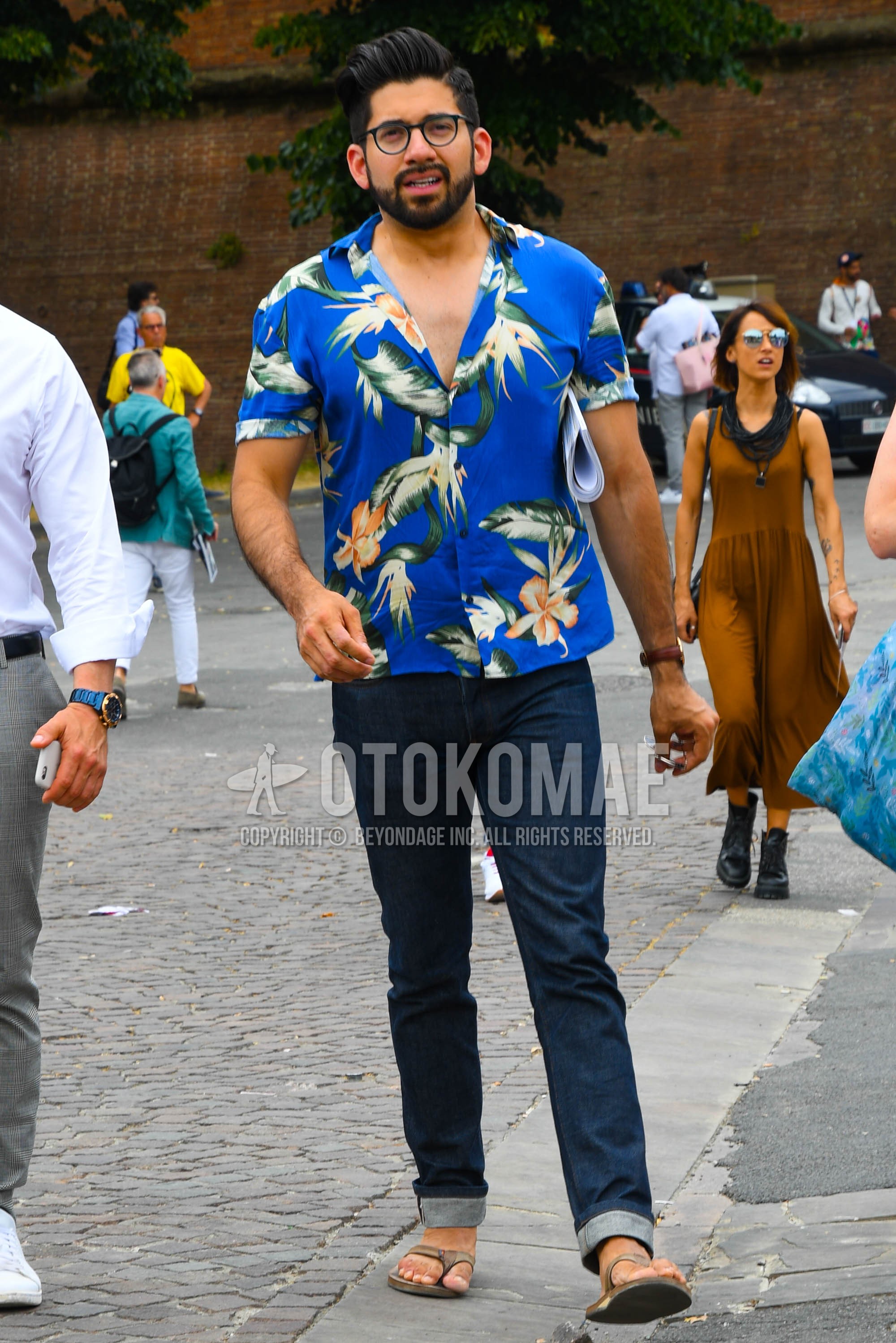 Men's summer outfit with plain glasses, blue botanical shirt, navy plain denim/jeans, beige flip flops sandals.