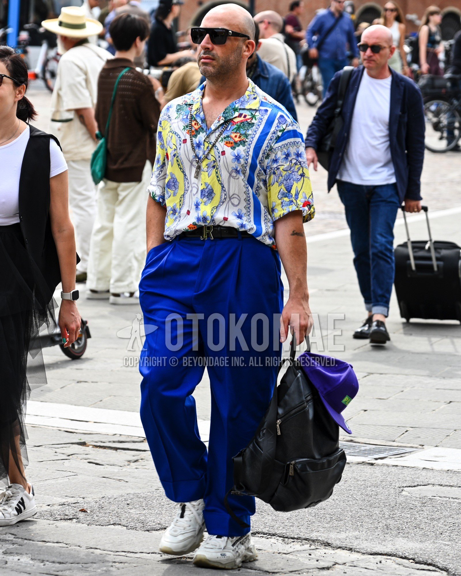 Men's spring summer outfit with black plain sunglasses, multi-color white whole pattern shirt, blue plain wide pants, white low-cut sneakers, purple plain backpack.