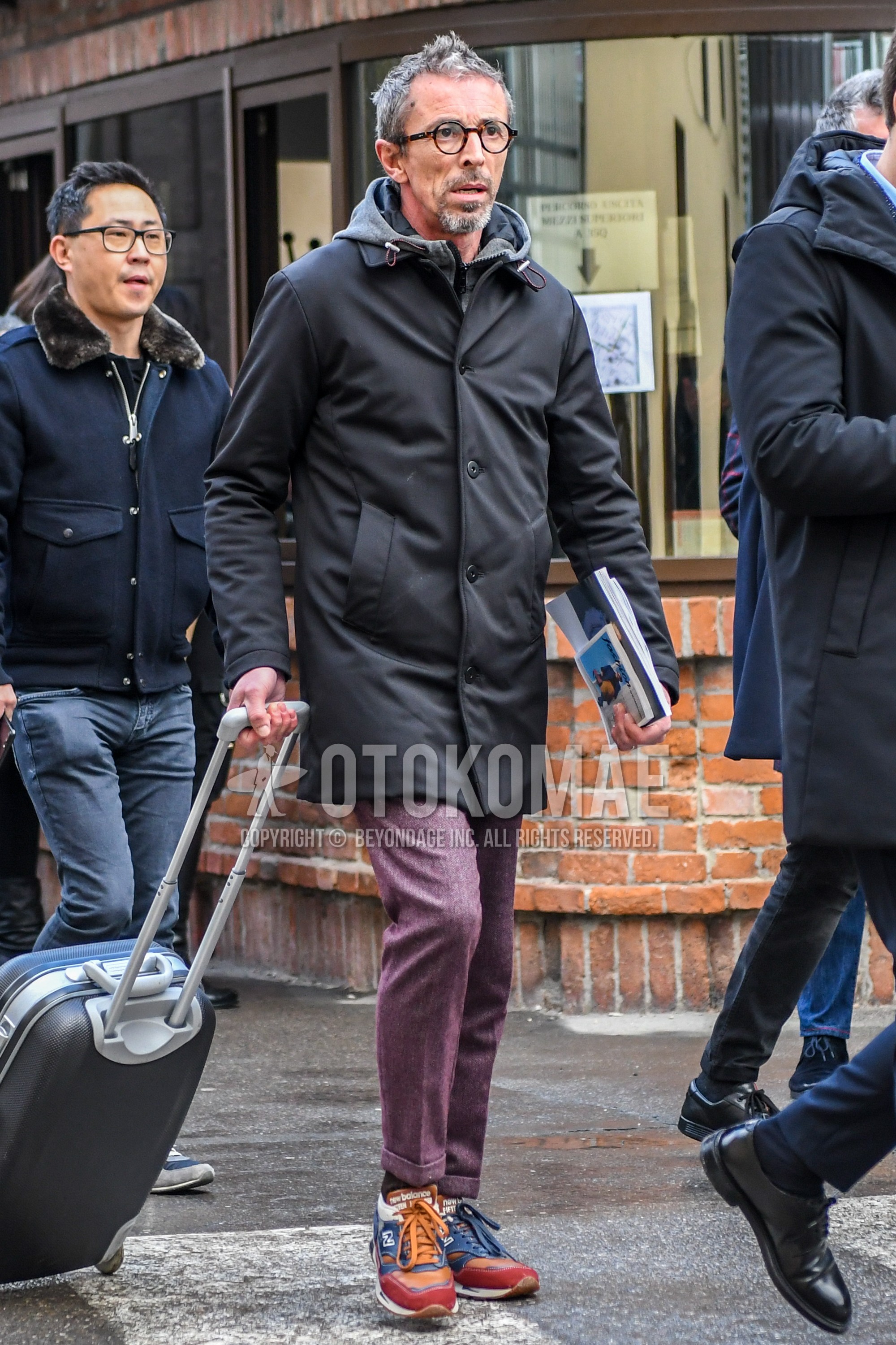 Men's winter outfit with brown tortoiseshell glasses, dark gray plain stenkarrer coat, gray plain hoodie, red plain slacks, brown plain socks, multi-color low-cut sneakers, black plain suitcase.