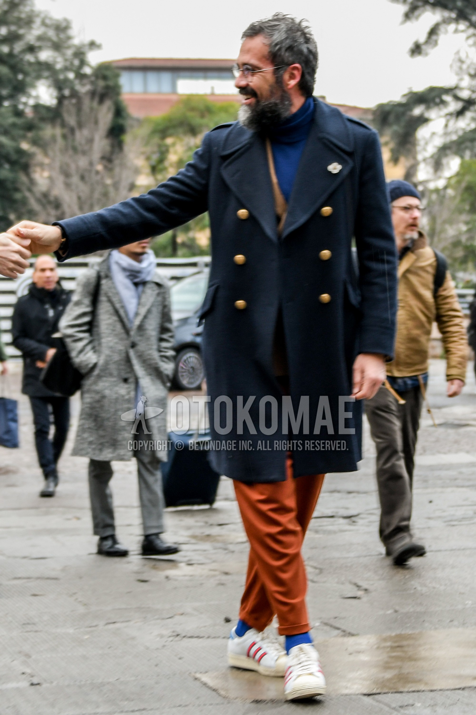 Men's autumn winter outfit with plain glasses, black plain ulster coat, beige plain tailored jacket, blue plain turtleneck knit, orange plain cotton pants, blue plain socks, white low-cut sneakers.