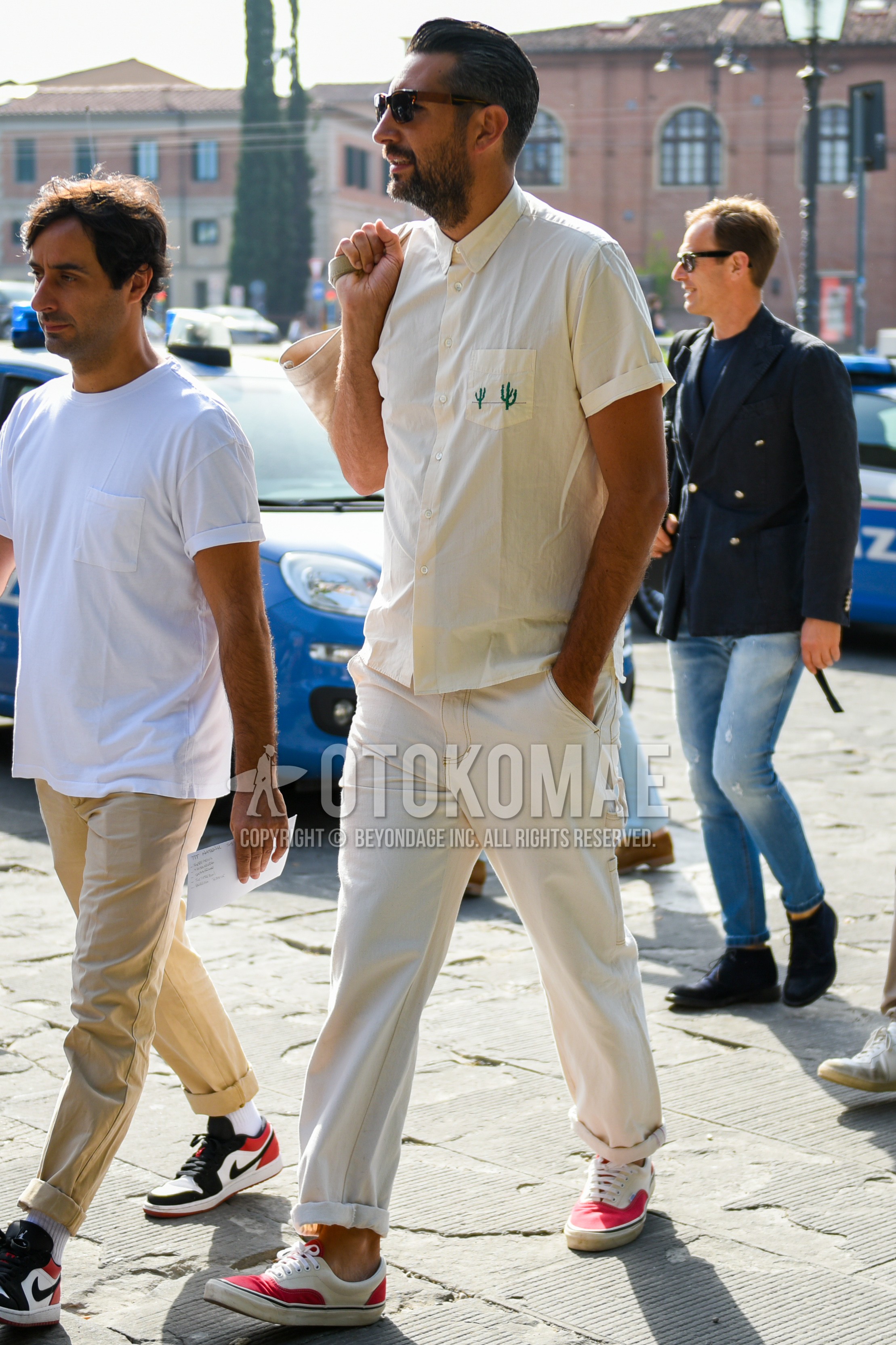 Men's spring summer outfit with brown tortoiseshell sunglasses, beige plain shirt, white plain cotton pants, red low-cut sneakers.