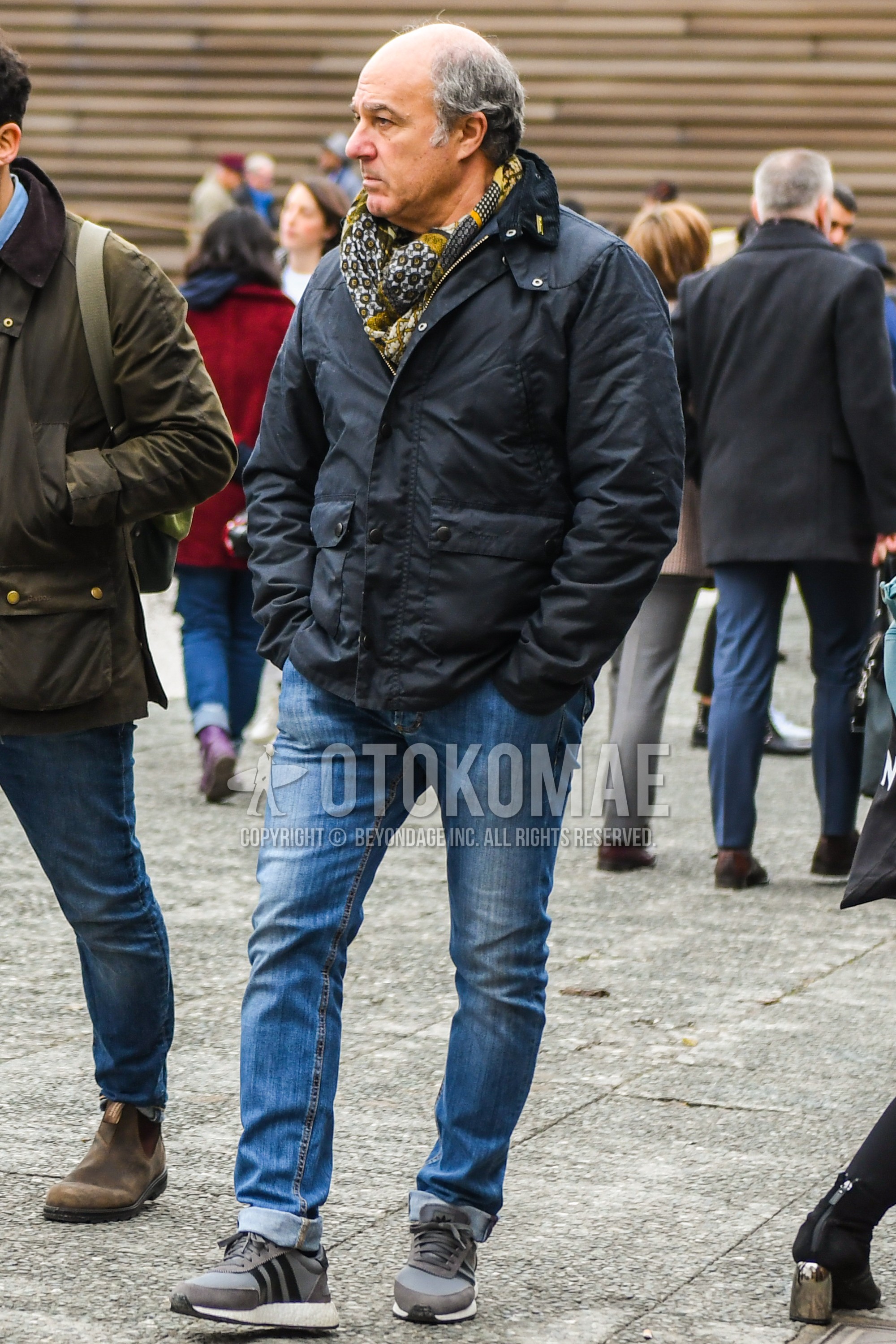 Men's winter outfit with yellow scarf scarf, black plain outerwear, light blue plain denim/jeans, gray low-cut sneakers.