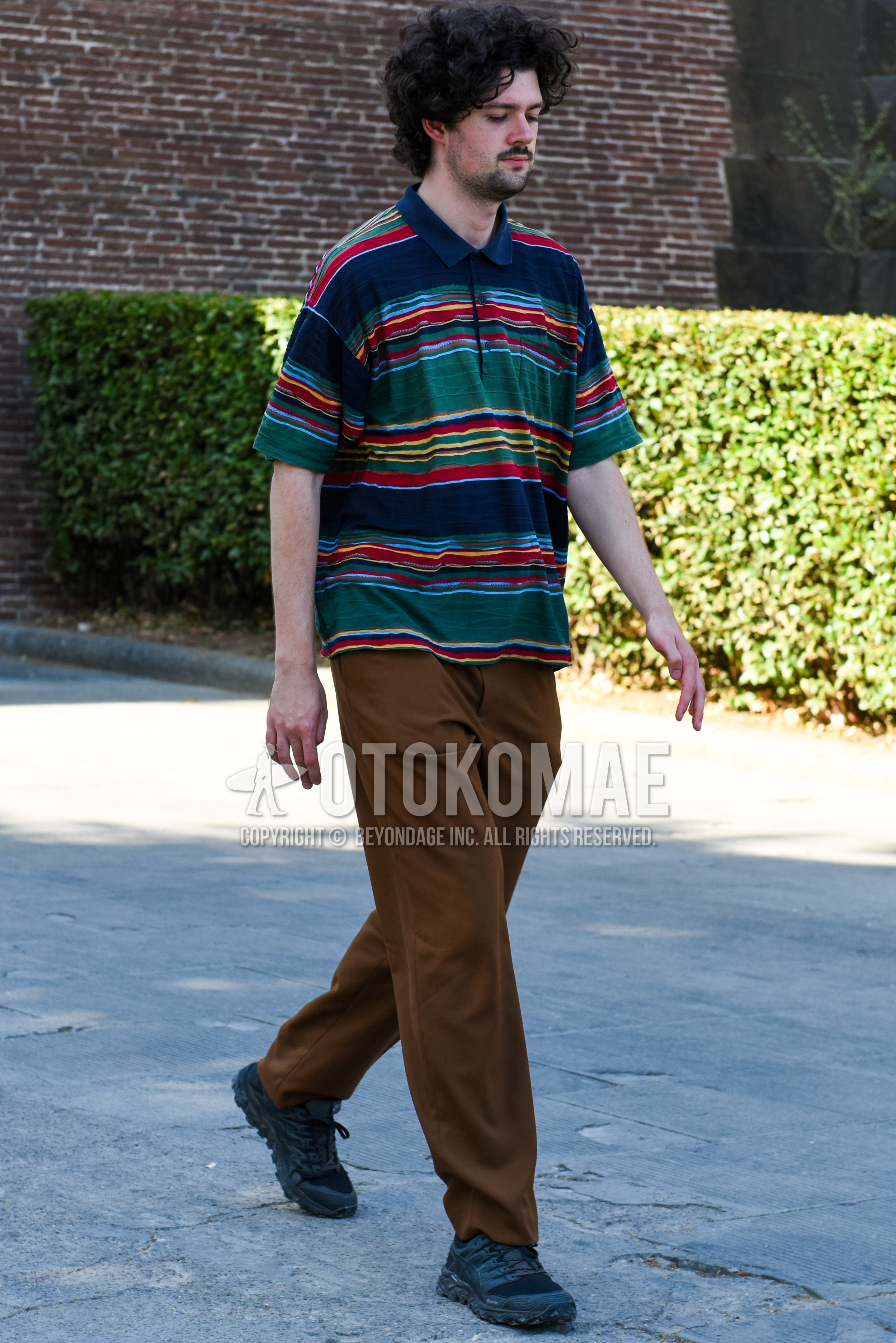 Men's spring summer outfit with multi-color horizontal stripes polo shirt, brown plain slacks, black low-cut sneakers.