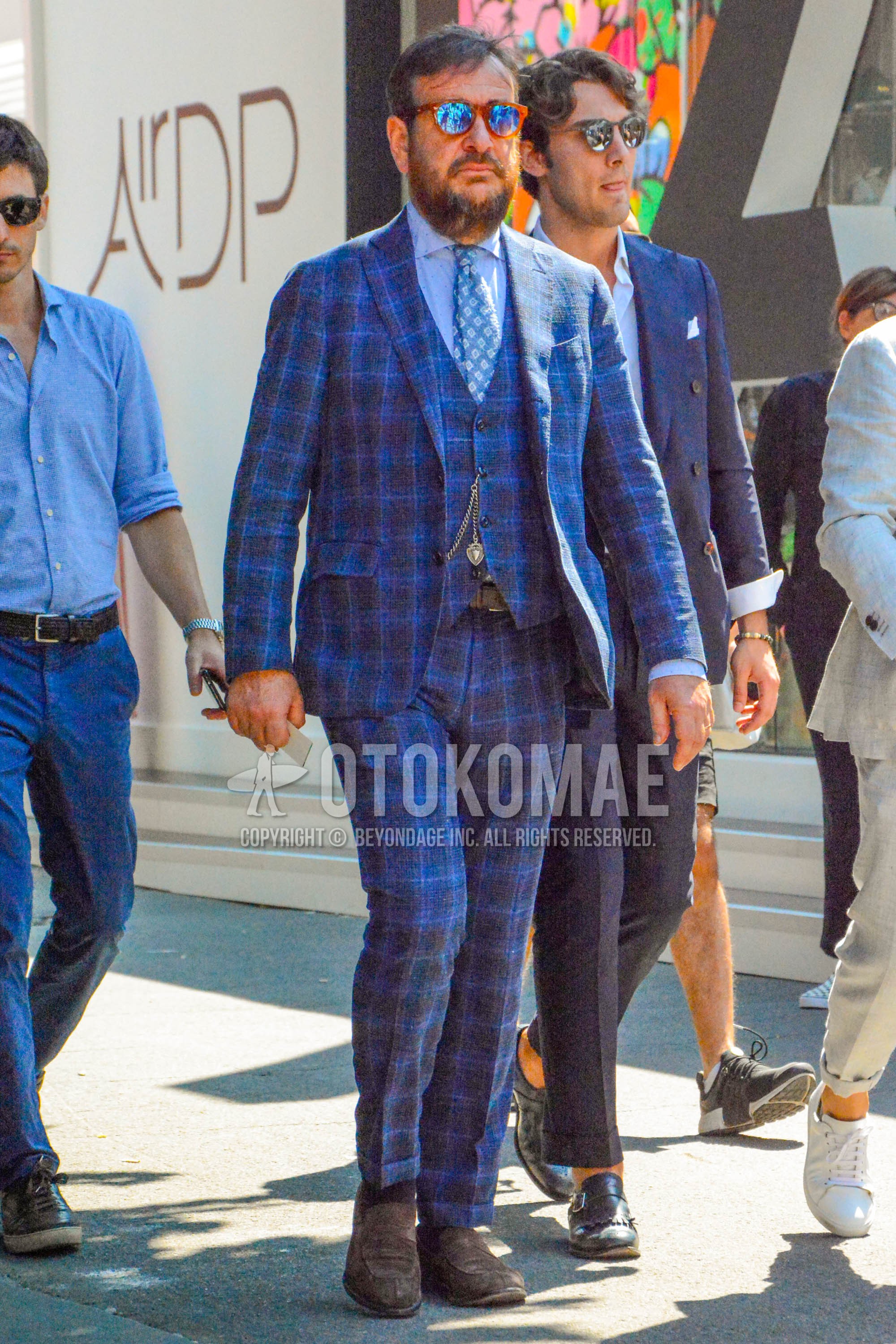 Men's spring autumn outfit with brown tortoiseshell sunglasses, blue dots shirt, black plain leather belt, brown coin loafers leather shoes, navy check three-piece suit, blue small crest necktie.