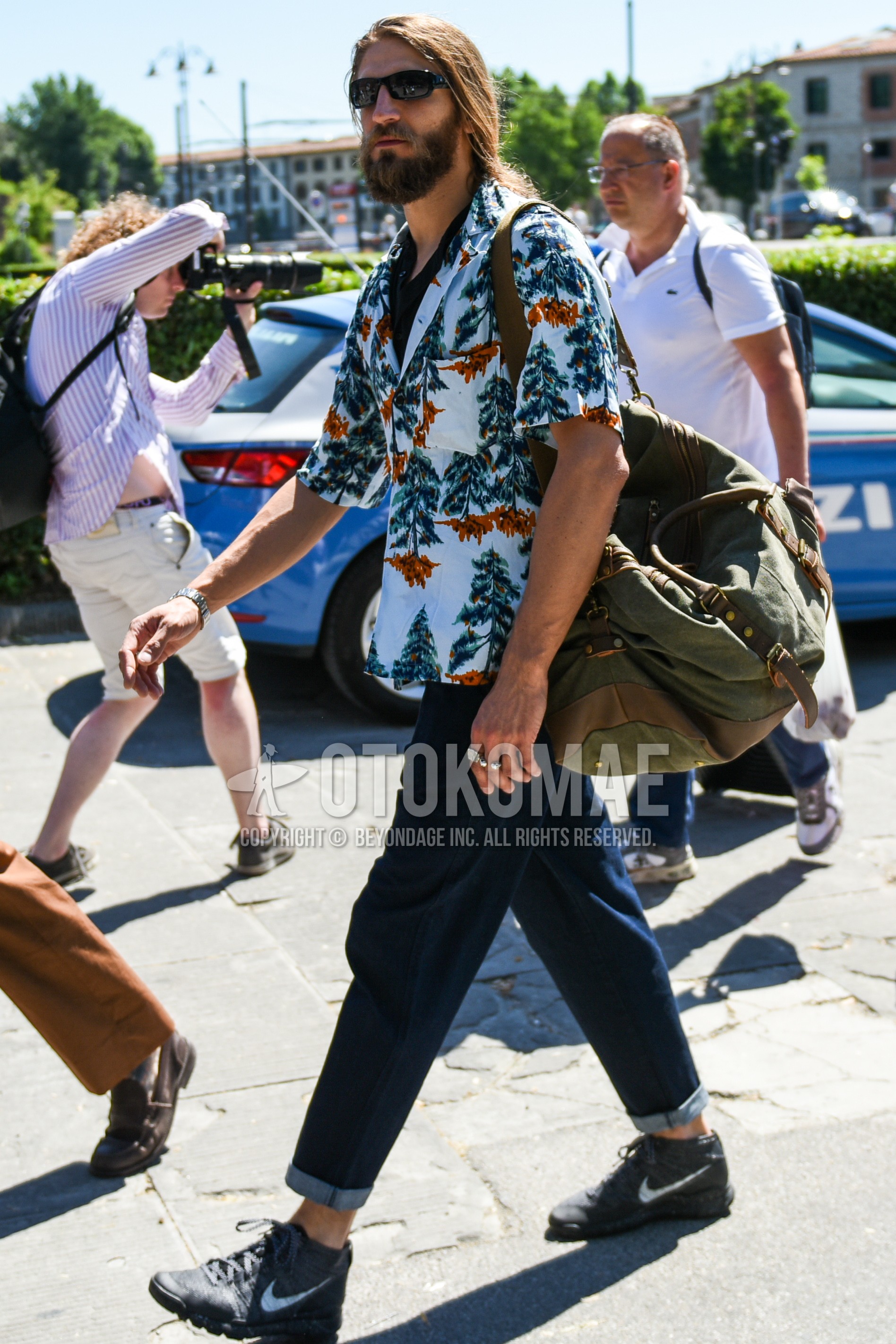 Men's spring summer outfit with black plain sunglasses, white botanical shirt, navy plain denim/jeans, dark gray high-cut sneakers, olive green plain shoulder bag.
