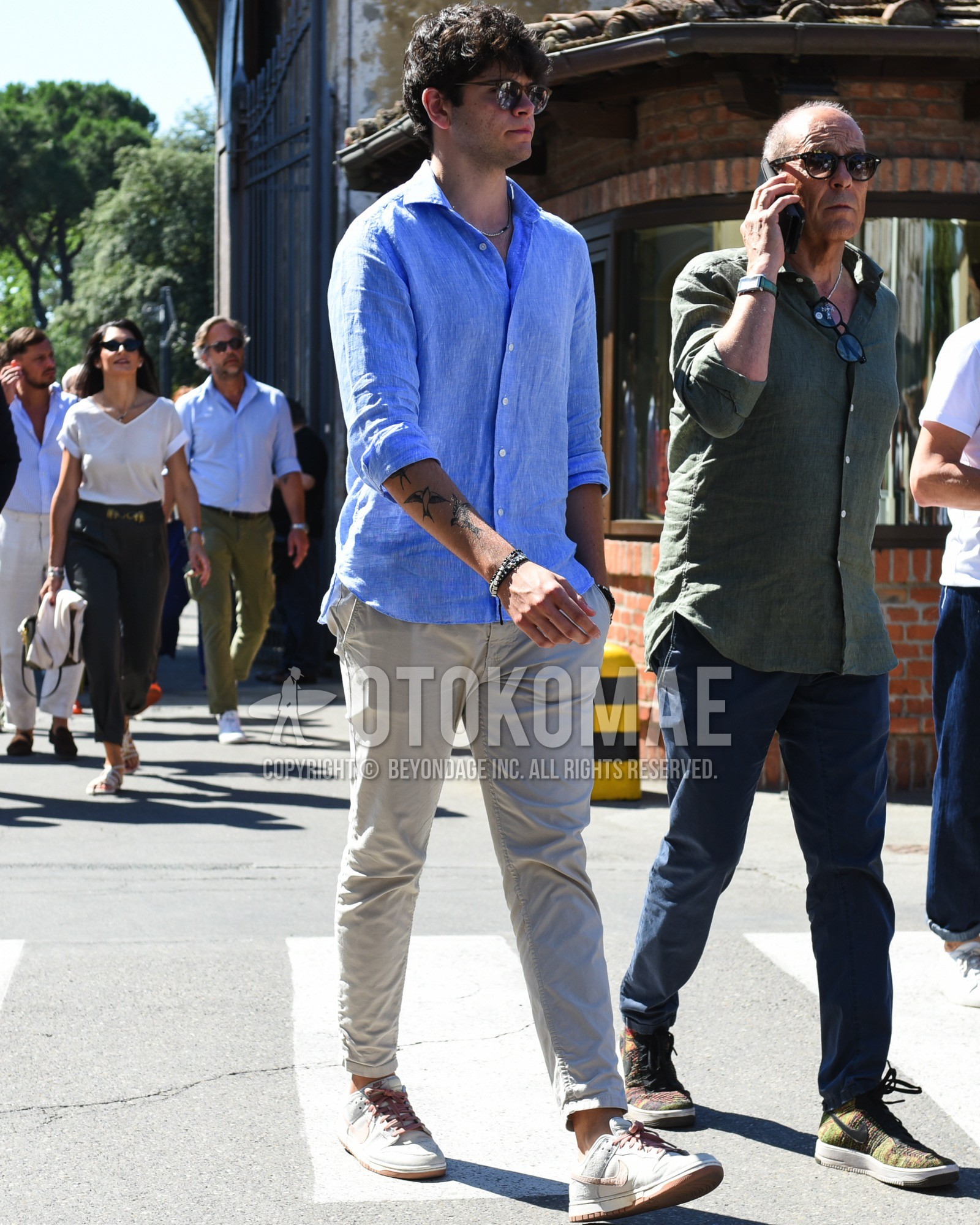 Men's spring summer outfit with brown plain sunglasses, blue plain shirt, gray plain chinos, gray white low-cut sneakers.