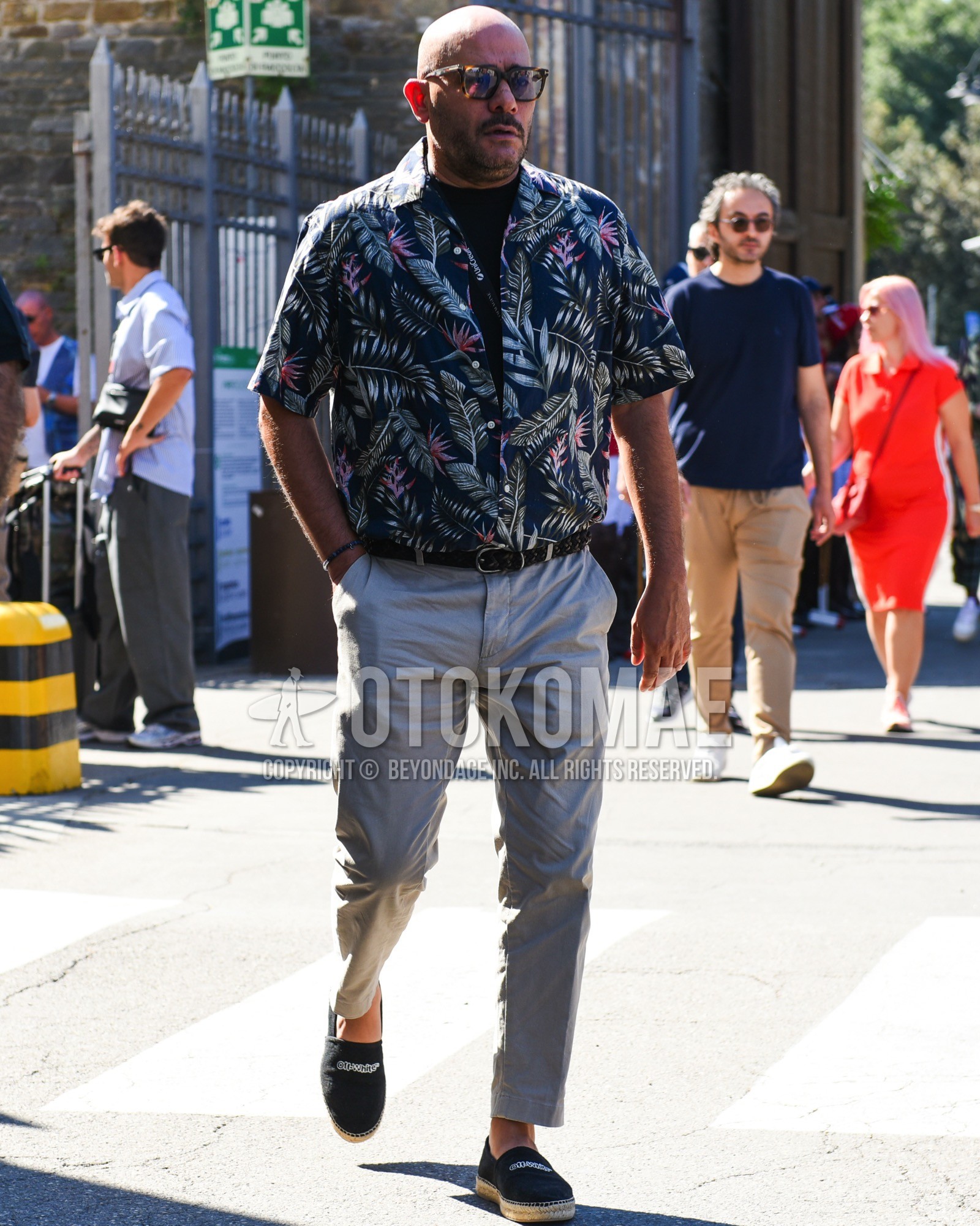 Men's spring summer outfit with brown tortoiseshell sunglasses, navy botanical shirt, black plain t-shirt, black plain braided belt, gray plain slacks, black  espadrille.
