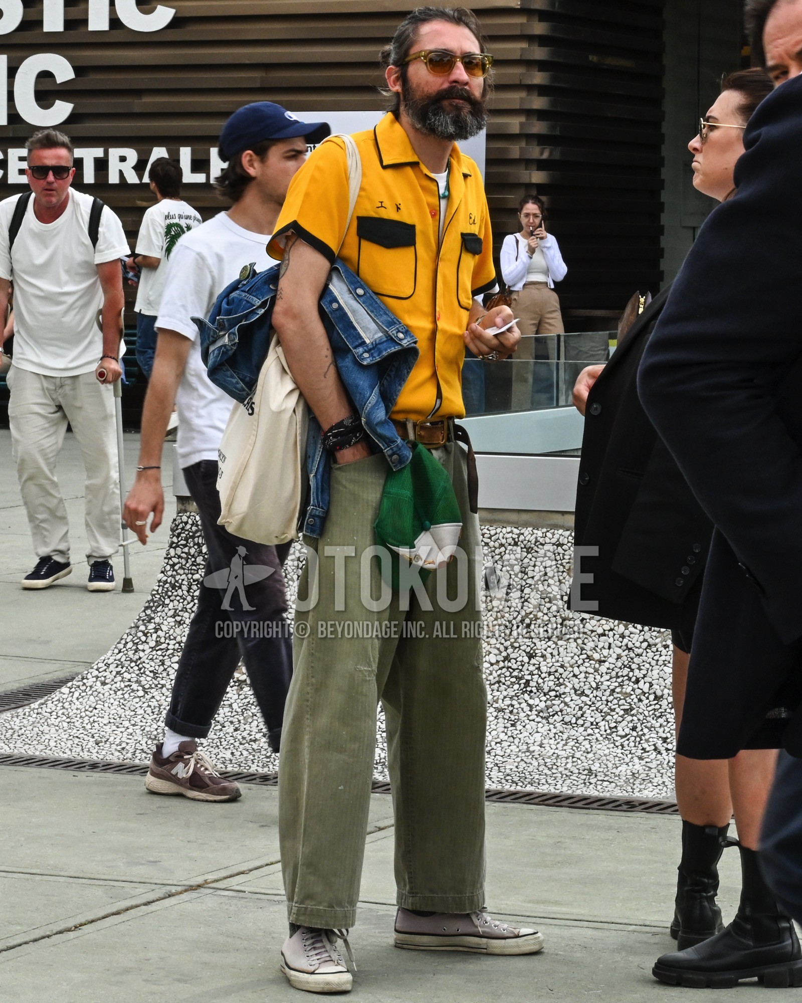 Men's spring summer outfit with brown plain sunglasses, yellow plain shirt, brown plain leather belt, olive green plain chinos, white low-cut sneakers, white plain tote bag.