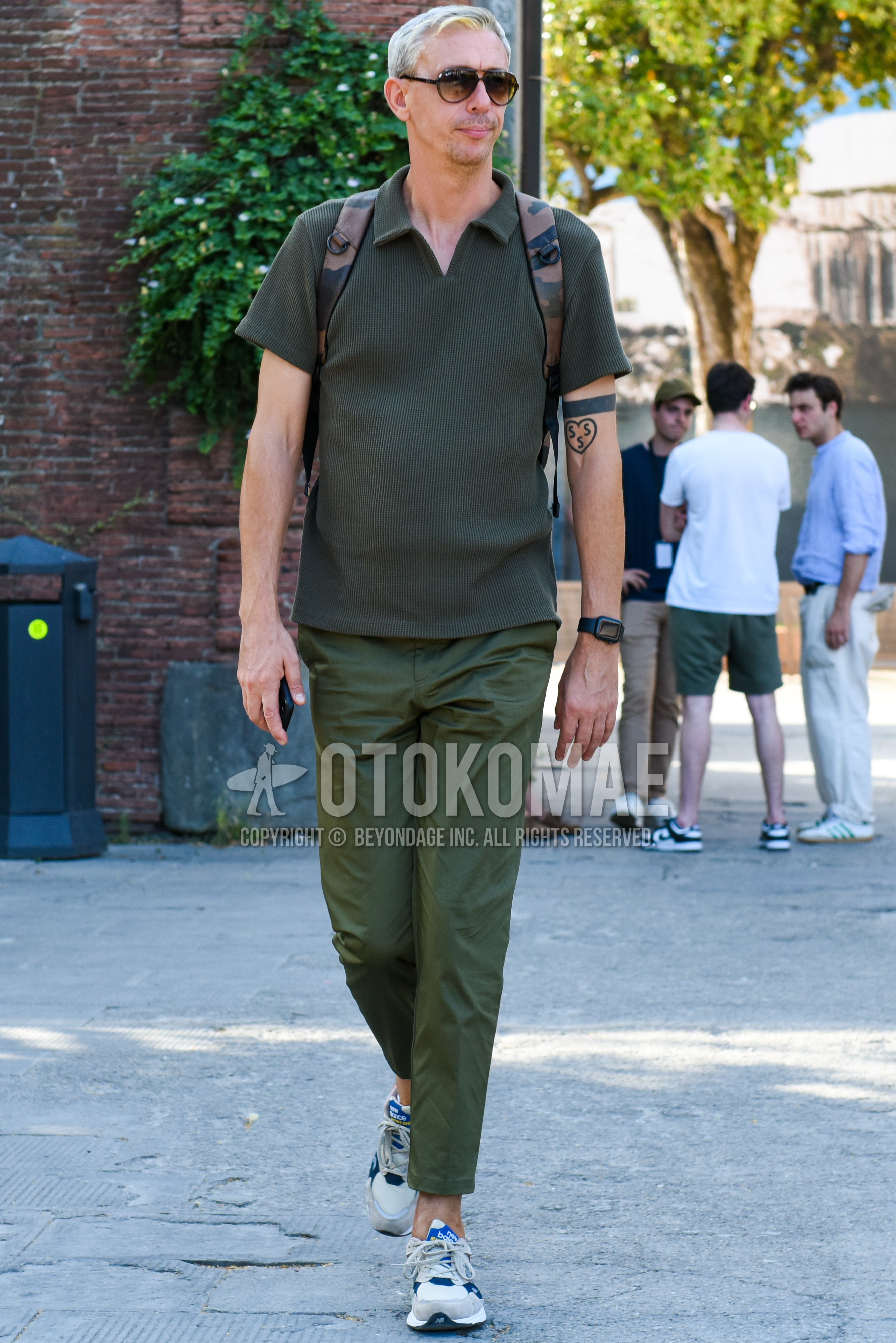 Men's spring summer autumn outfit with brown plain sunglasses, green plain t-shirt, green plain cotton pants, white low-cut sneakers, olive green brown camouflage backpack.