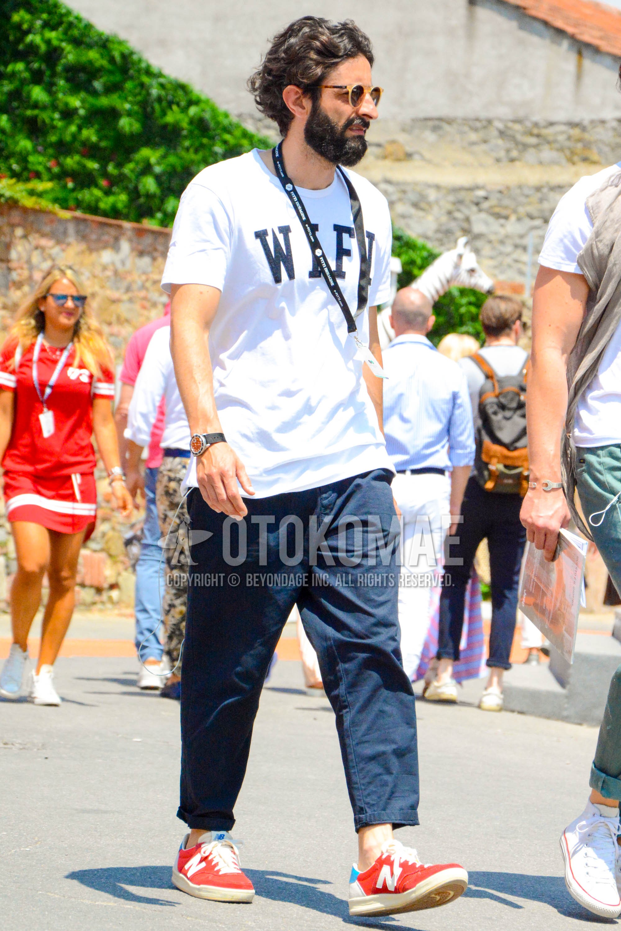 Men's summer outfit with plain sunglasses, white graphic t-shirt, navy plain ankle pants, red low-cut sneakers.