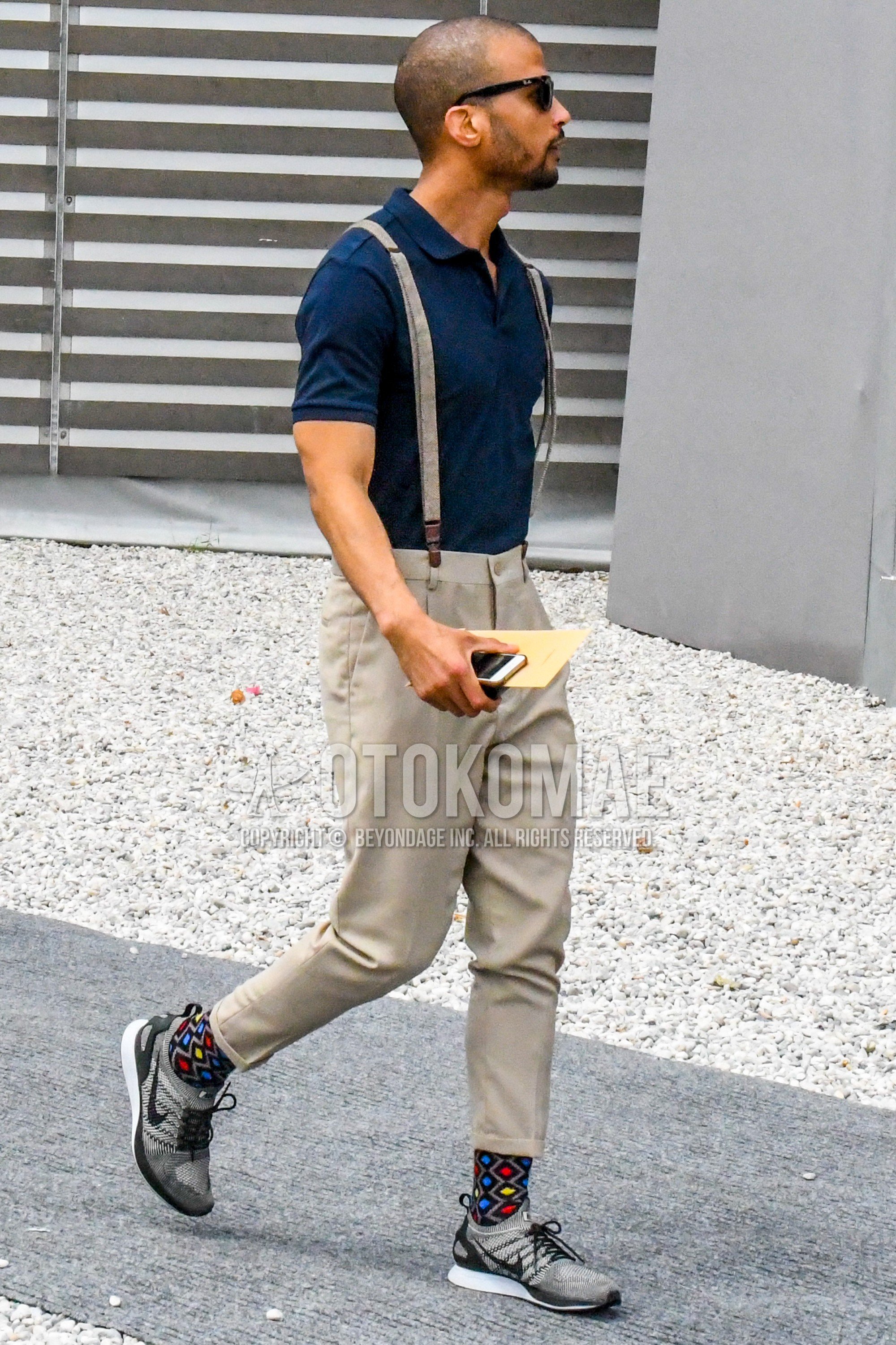 Men's summer outfit with plain sunglasses, navy plain polo shirt, beige plain suspenders, beige plain cropped pants, multi-color socks socks, gray low-cut sneakers.