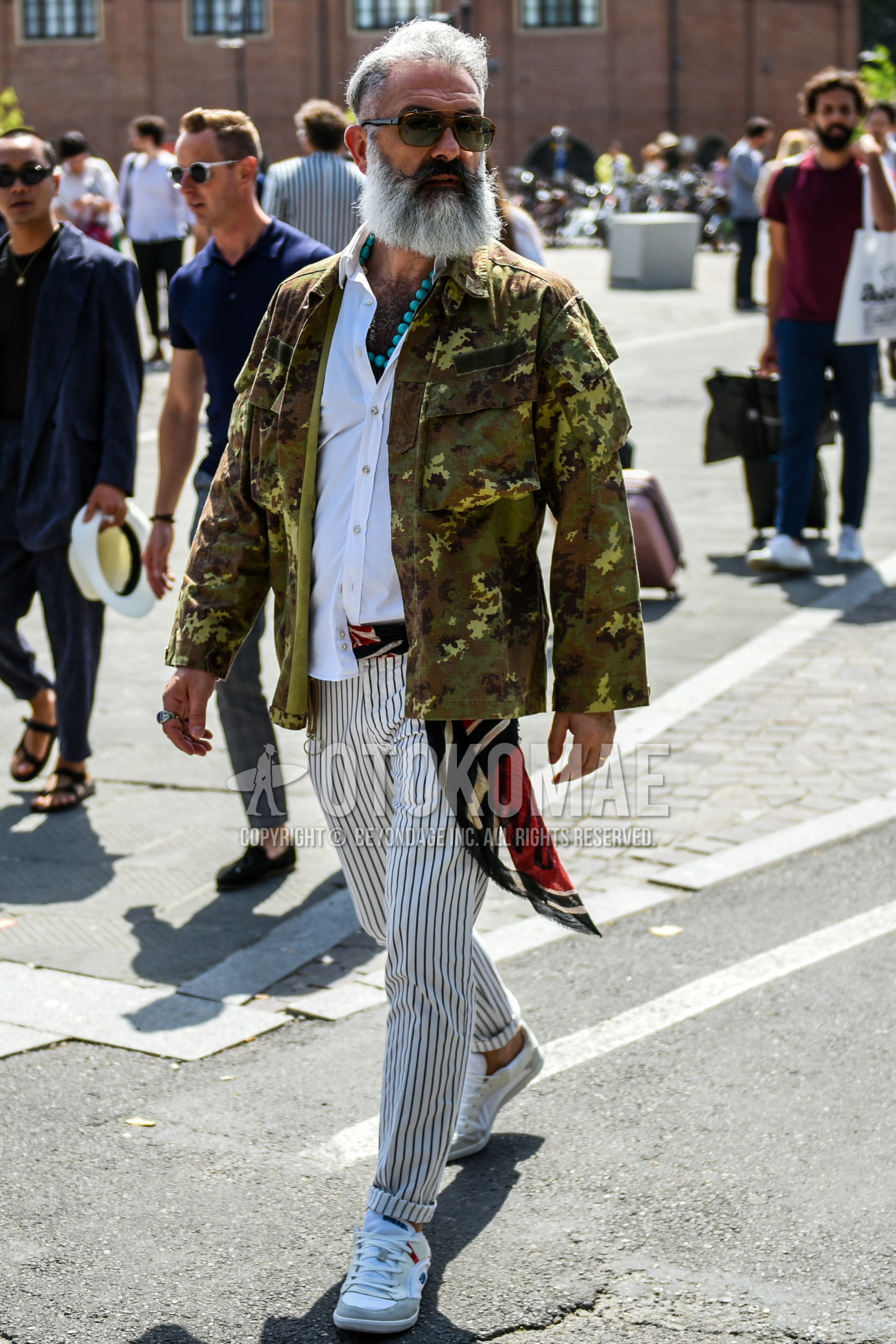 Men's spring autumn outfit with brown plain sunglasses, multi-color scarf scarf, green camouflage shirt jacket, white plain shirt, multi-color belt tape belt, white stripes slacks, white low-cut sneakers.