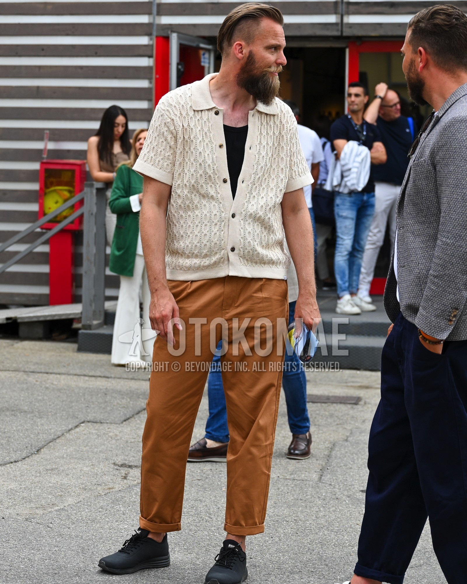 Men's spring summer outfit with beige plain cardigan, black plain t-shirt, brown plain chinos, black low-cut sneakers.