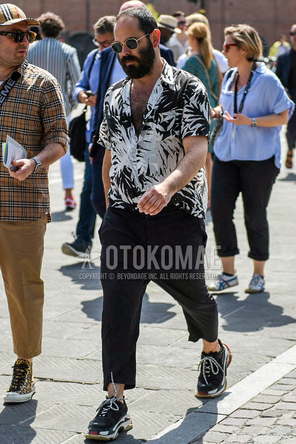 Men's summer outfit with silver plain sunglasses, black white botanical shirt, black plain ankle pants, black low-cut sneakers.