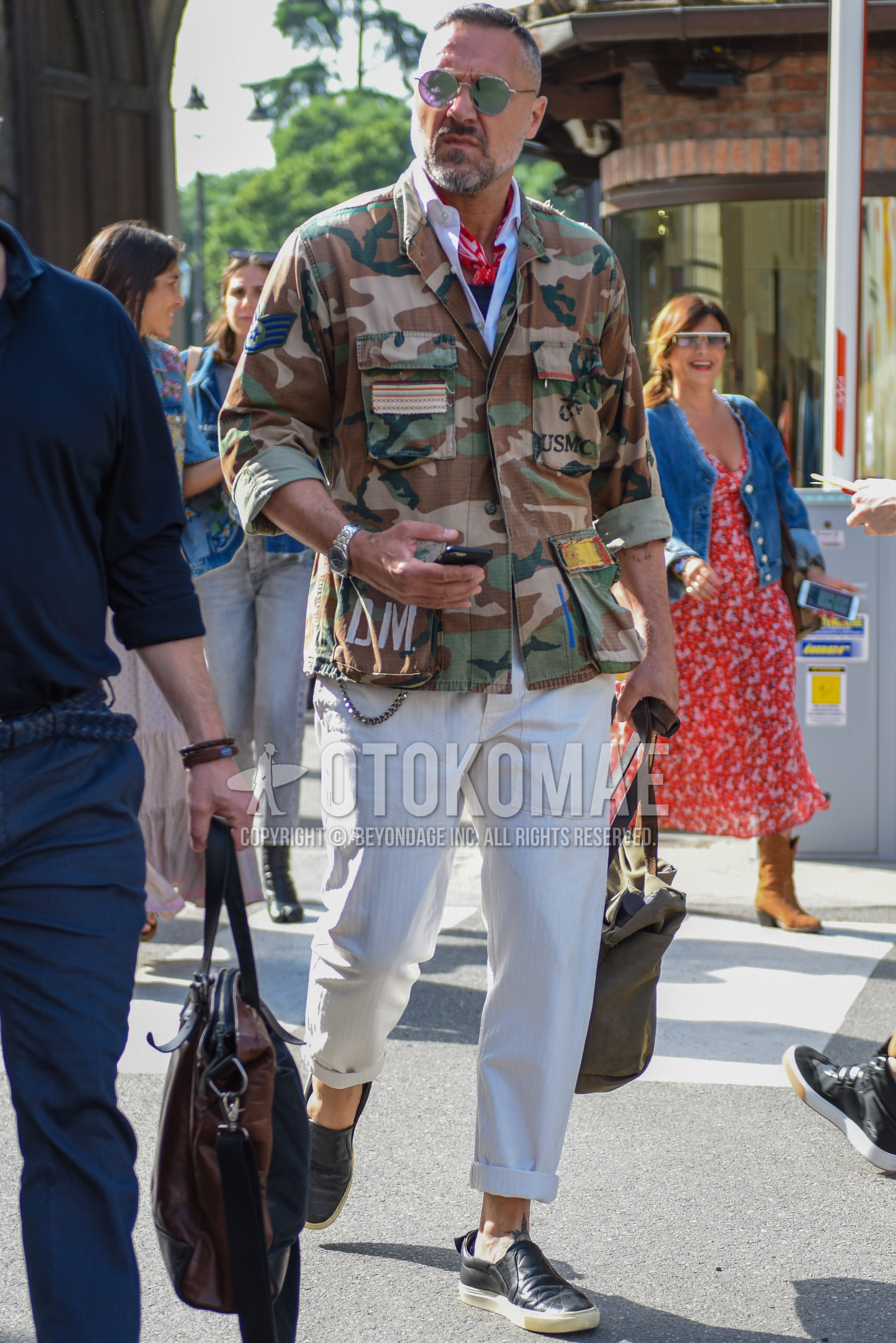 Men's spring autumn outfit with silver plain sunglasses, red scarf bandana/neckerchief, olive green camouflage shirt jacket, white plain shirt, beige plain cotton pants, black slip-on sneakers.
