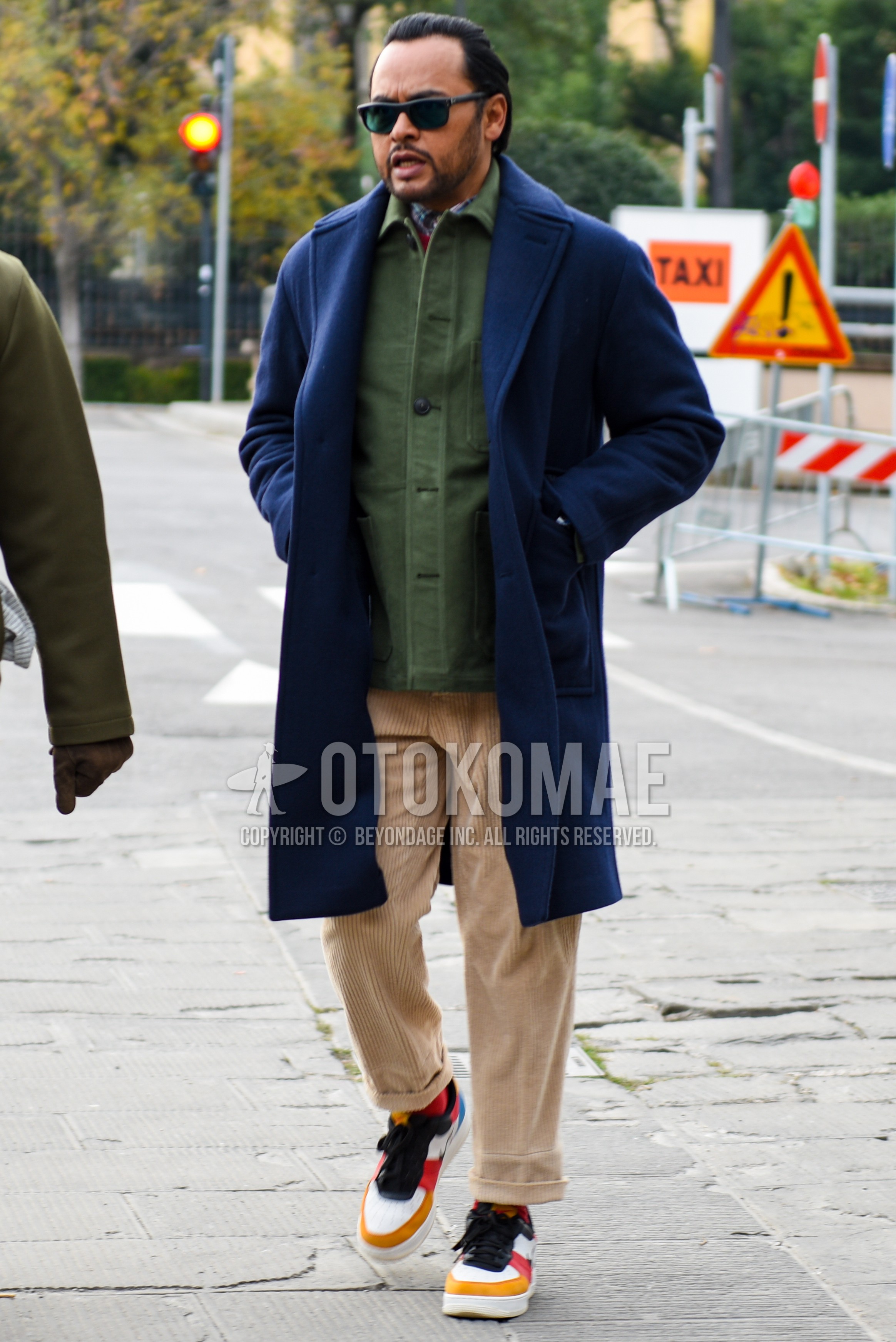 Men's autumn winter outfit with black plain sunglasses, navy plain chester coat, olive green plain shirt jacket, beige plain winter pants (corduroy,velour), red plain socks, multi-color low-cut sneakers.