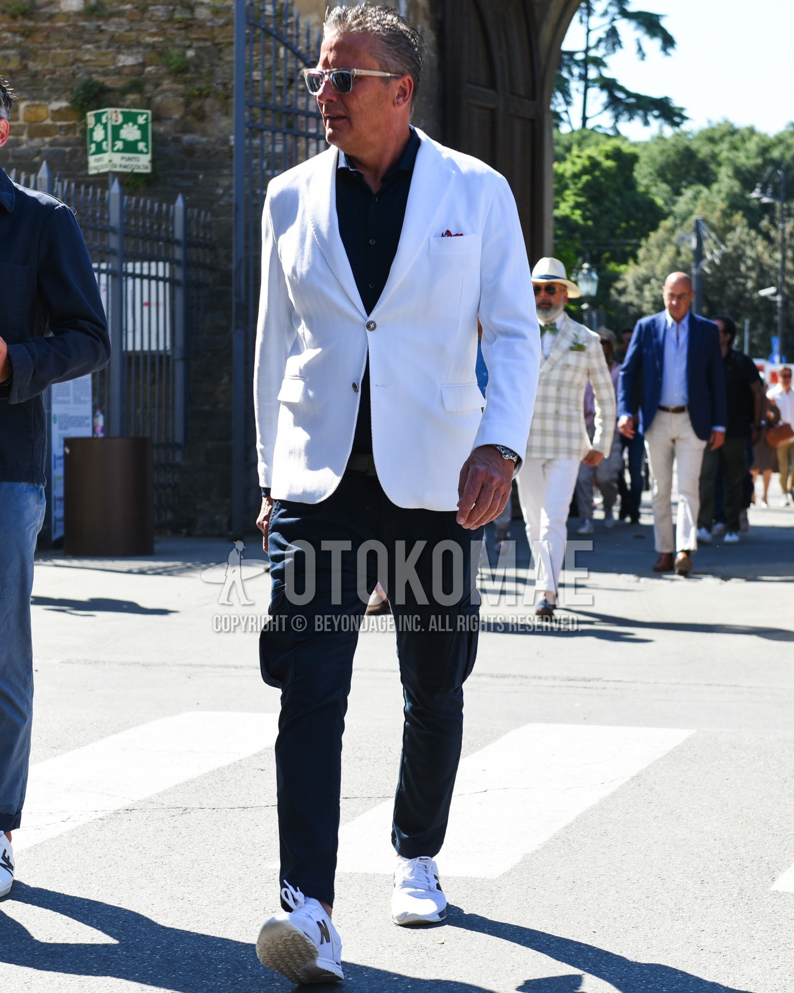 Men's spring summer outfit with clear plain sunglasses, white plain tailored jacket, navy plain shirt, navy plain cargo pants, white low-cut sneakers.