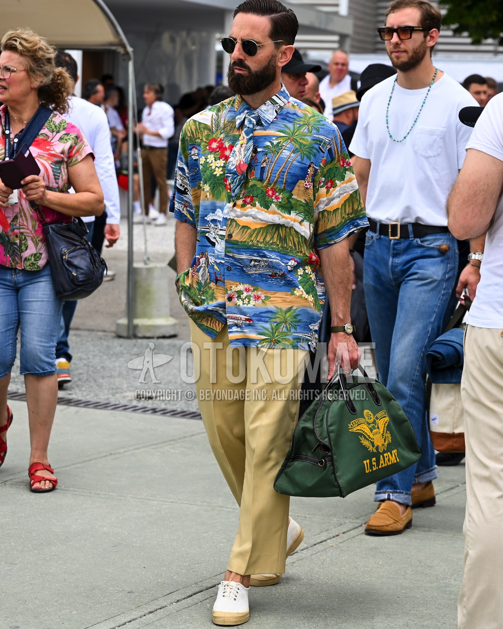 Men's spring summer outfit with black plain sunglasses, blue whole pattern shirt, beige plain chinos, white low-cut sneakers, olive green bag boston bag.