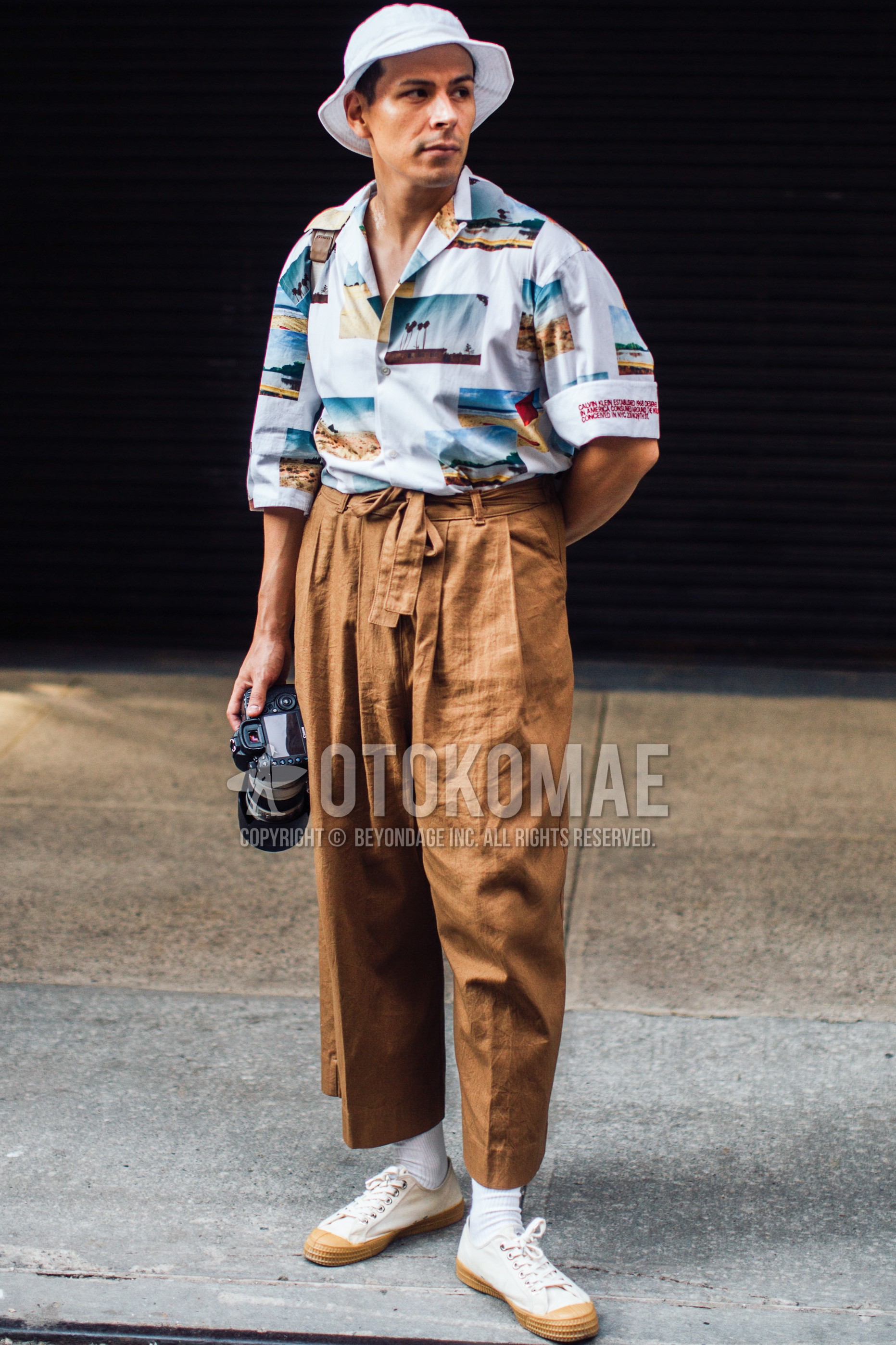 Men's spring summer outfit with white plain cap, white graphic shirt, brown beige plain cropped pants, white plain socks, white sneakers, white plain shoulder bag.