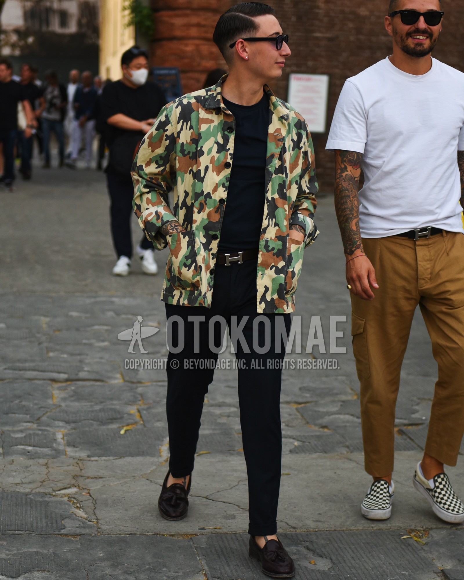 Men's spring summer outfit with black plain sunglasses, beige camouflage shirt jacket, black plain t-shirt, black plain leather belt, black plain slacks, brown tassel loafers leather shoes.
