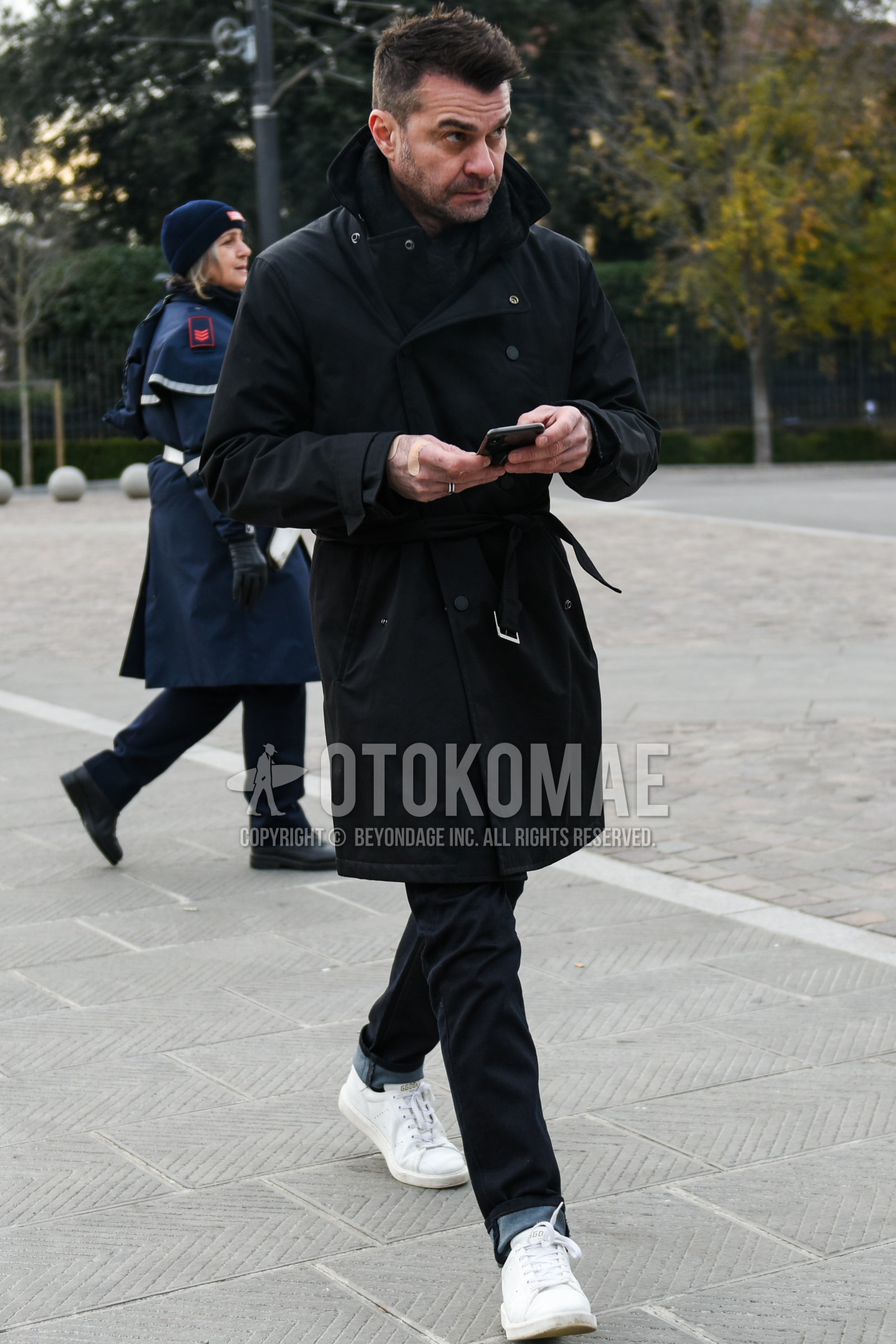 Men's autumn winter outfit with black plain scarf, black plain belted coat, black plain denim/jeans, white low-cut sneakers.