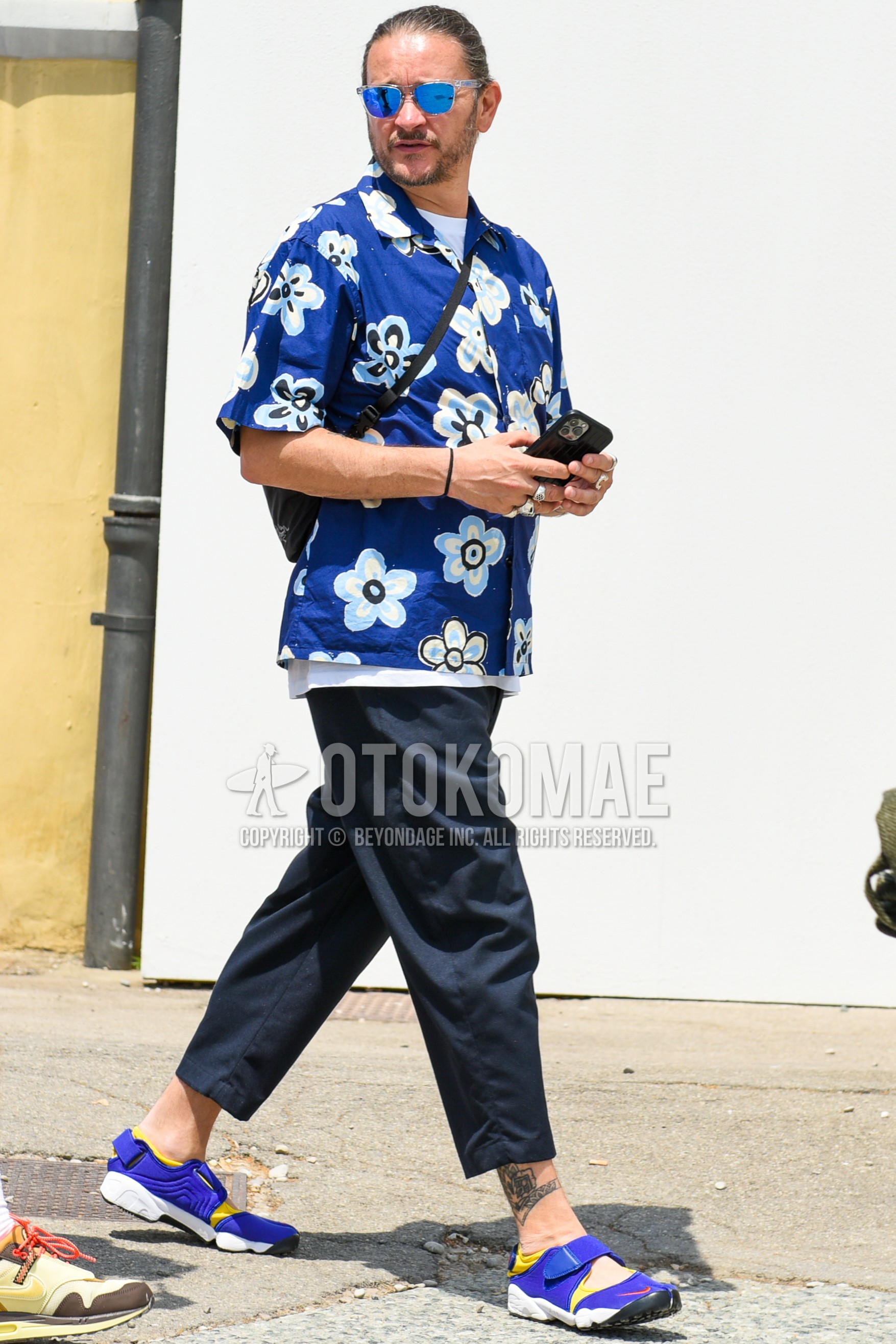 Men's spring summer outfit with clear plain sunglasses, blue tops/innerwear shirt, white plain t-shirt, navy plain ankle pants, navy plain cropped pants, blue sport sandals, black plain body bag.