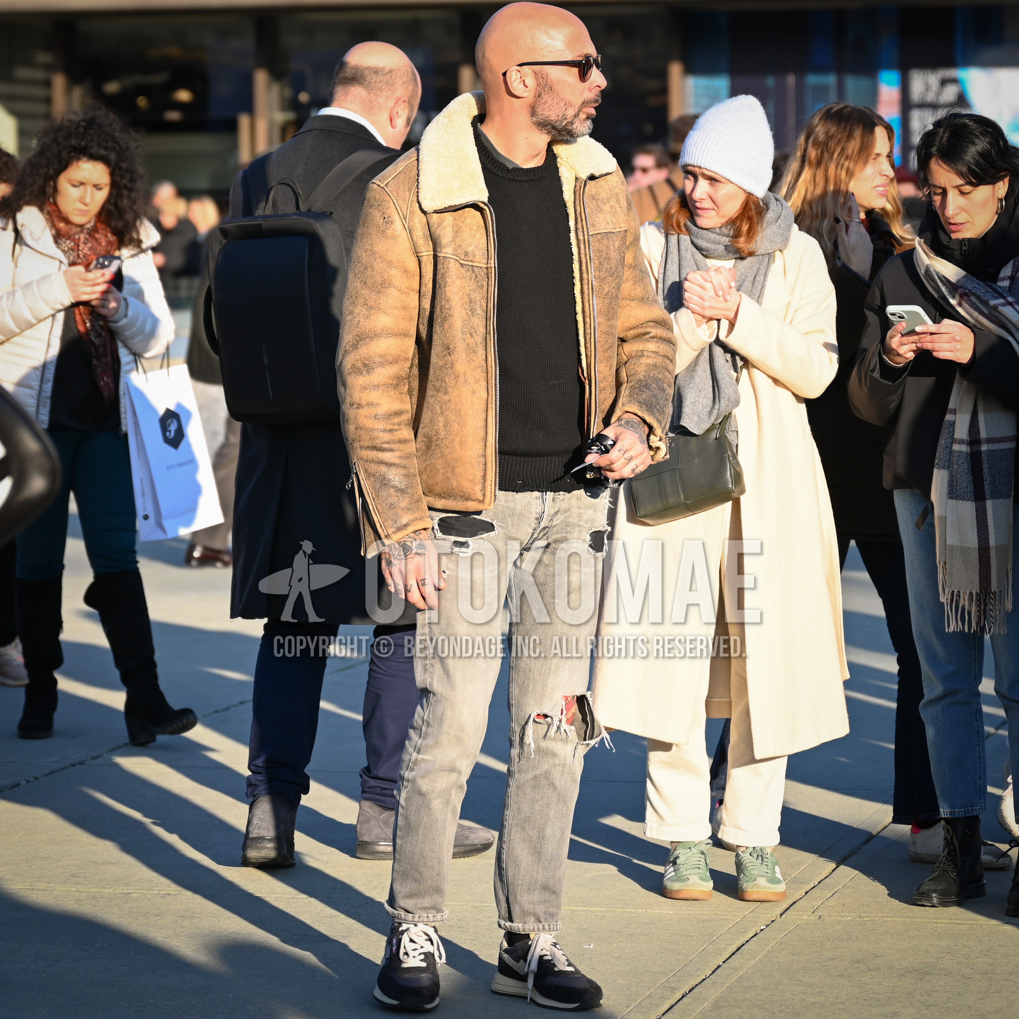 Men's autumn winter outfit with black plain sunglasses, brown beige outerwear, black plain sweater, gray bottoms damaged jeans, black plain socks, navy low-cut sneakers.