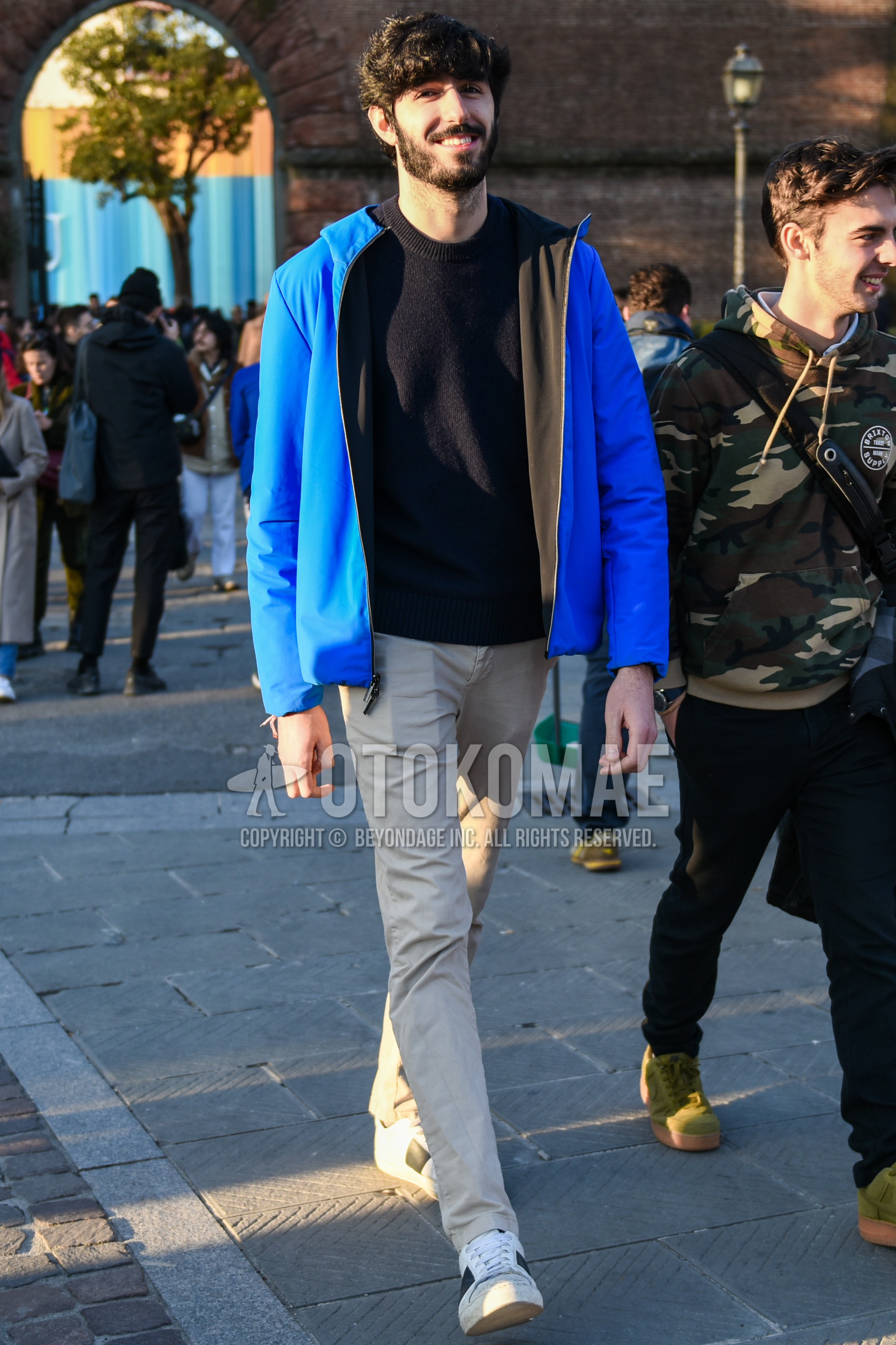 Men's autumn winter outfit with blue plain hooded coat, black plain sweater, beige plain chinos, white low-cut sneakers.