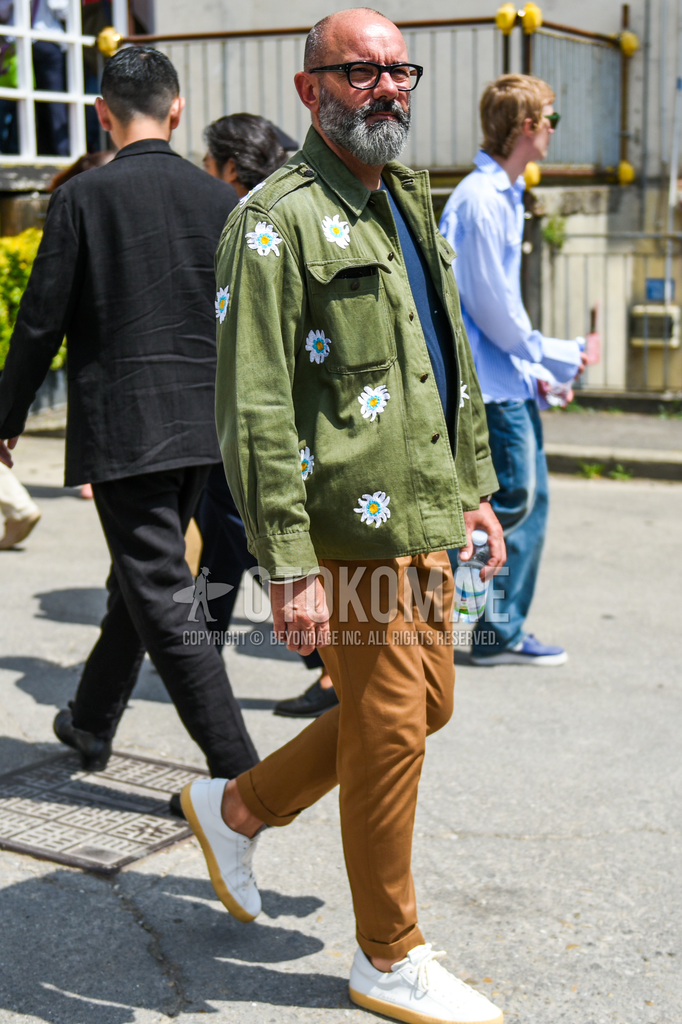 Men's spring autumn outfit with black plain glasses, olive green plain shirt jacket, navy plain t-shirt, brown plain slacks, white low-cut sneakers.