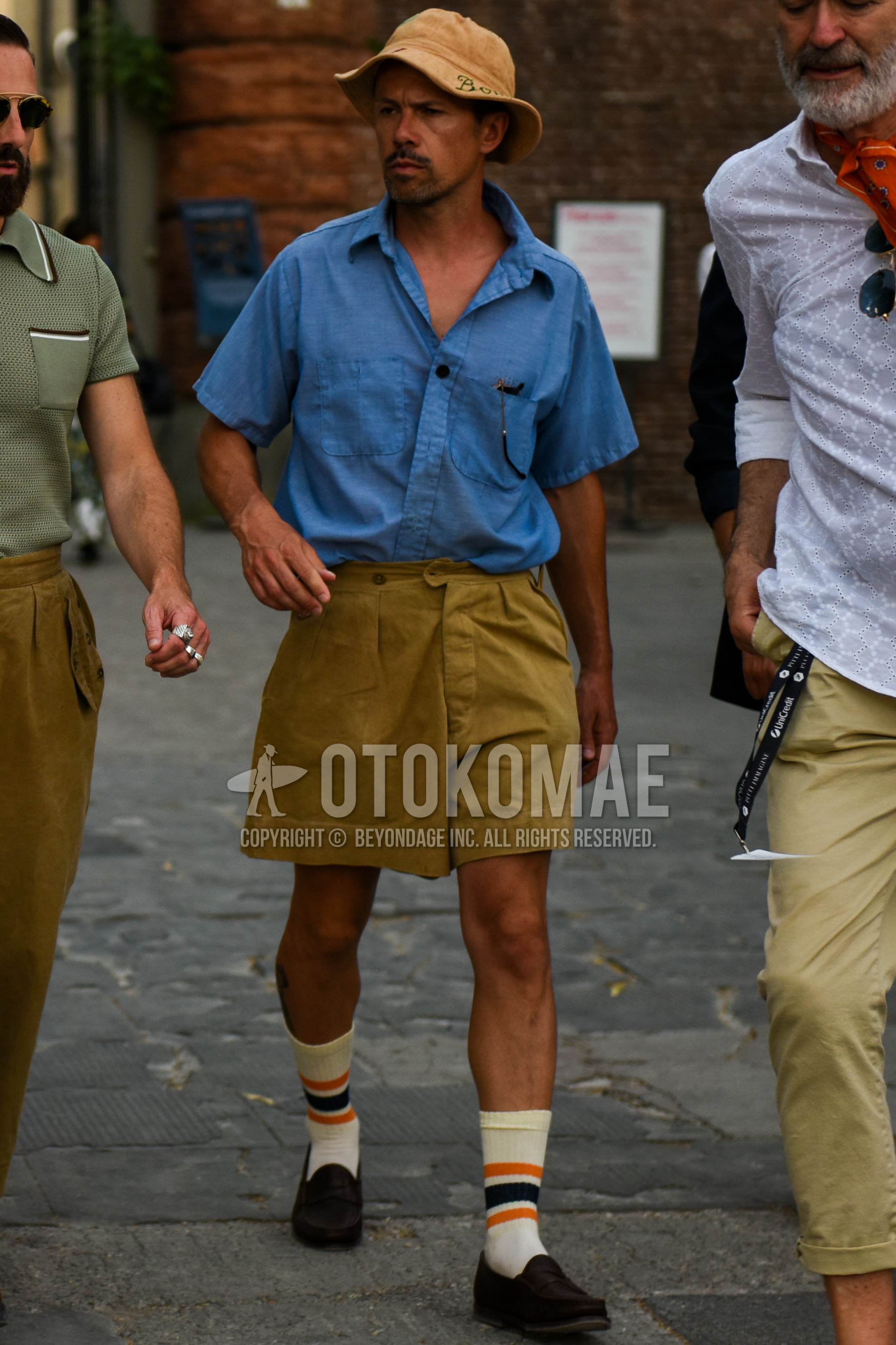 Men's spring summer outfit with brown plain bucket hat, blue plain shirt, beige olive green plain short pants, white horizontal stripes socks, black coin loafers leather shoes.