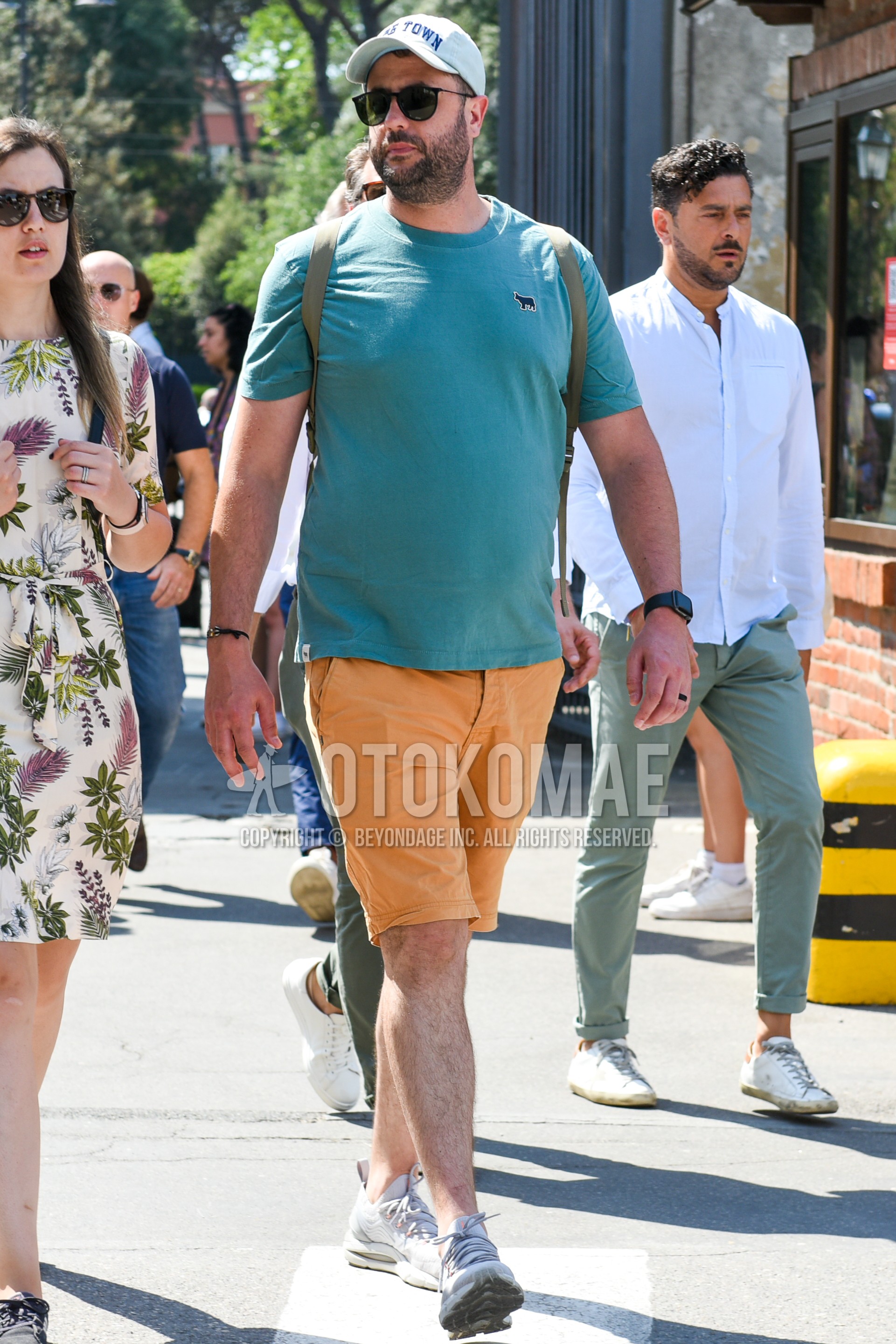 Men's spring summer outfit with white one point baseball cap, green plain t-shirt, orange plain short pants, gray low-cut sneakers, green plain backpack.