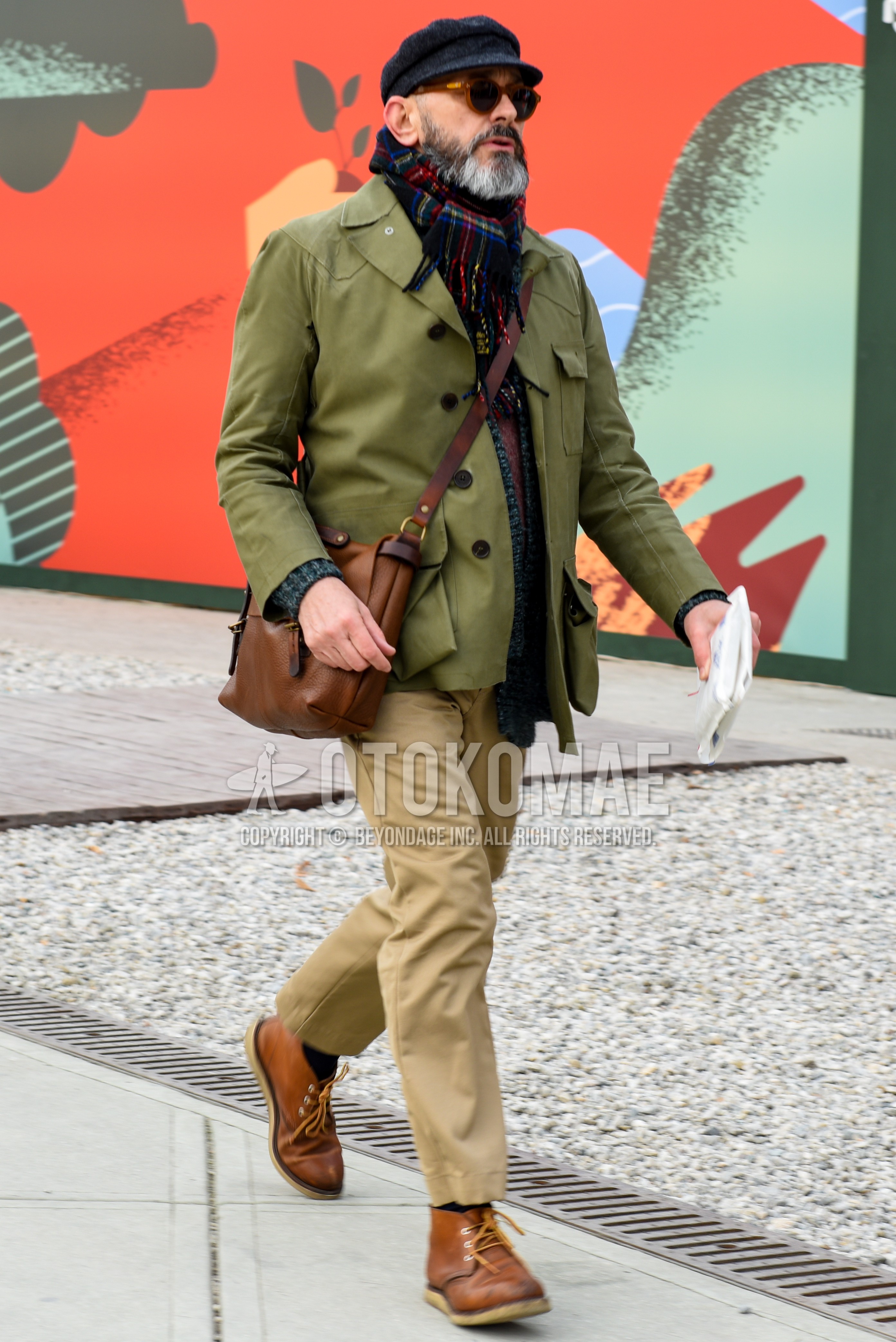Men's autumn winter outfit with silver plain cap, multi-color check scarf, olive green plain outerwear, beige plain chinos, beige plain cropped pants, navy plain socks, brown chukka boots, brown plain shoulder bag.