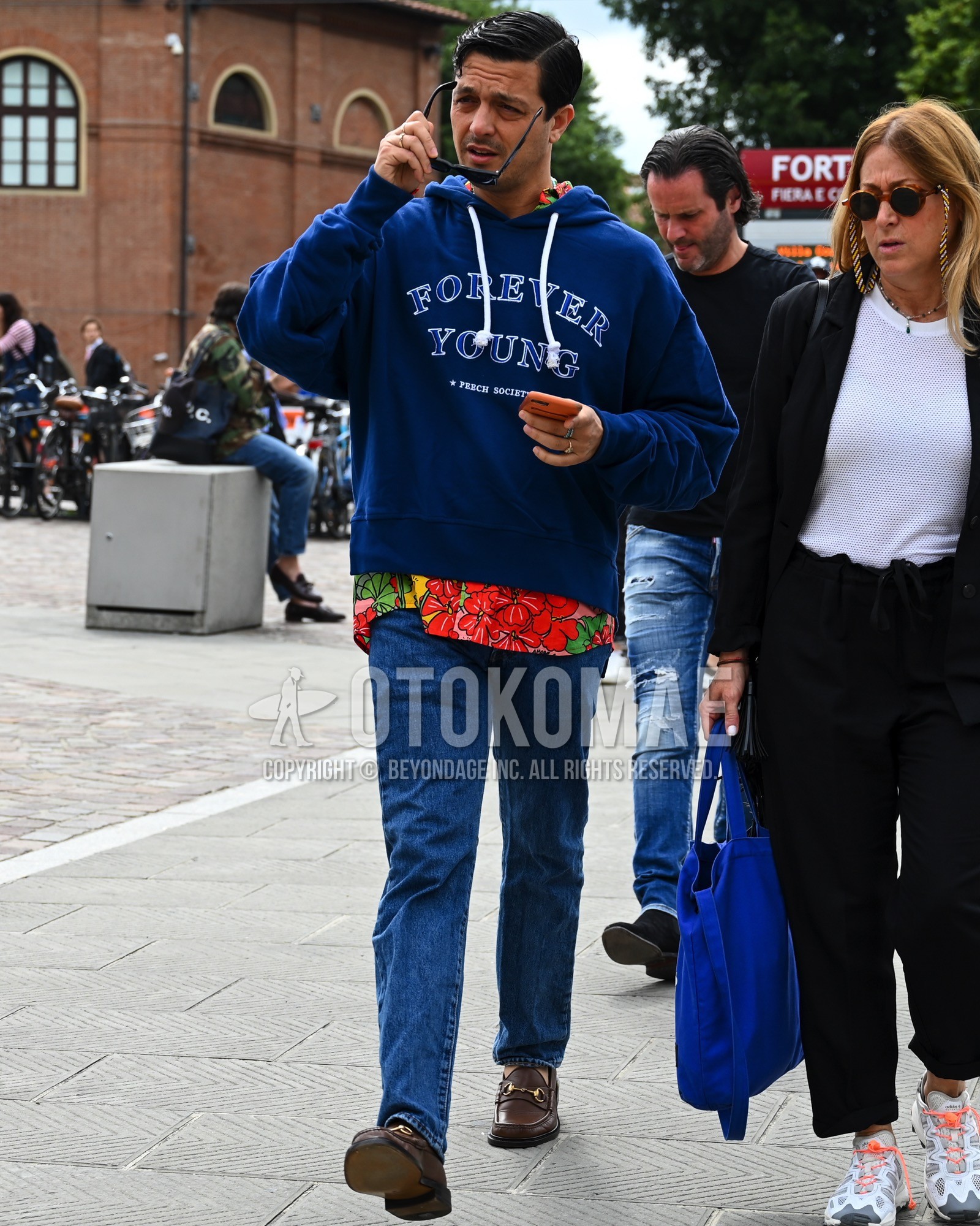 Men's spring summer autumn outfit with black plain sunglasses, blue lettered hoodie, red whole pattern shirt, blue plain denim/jeans, brown bit loafers leather shoes.
