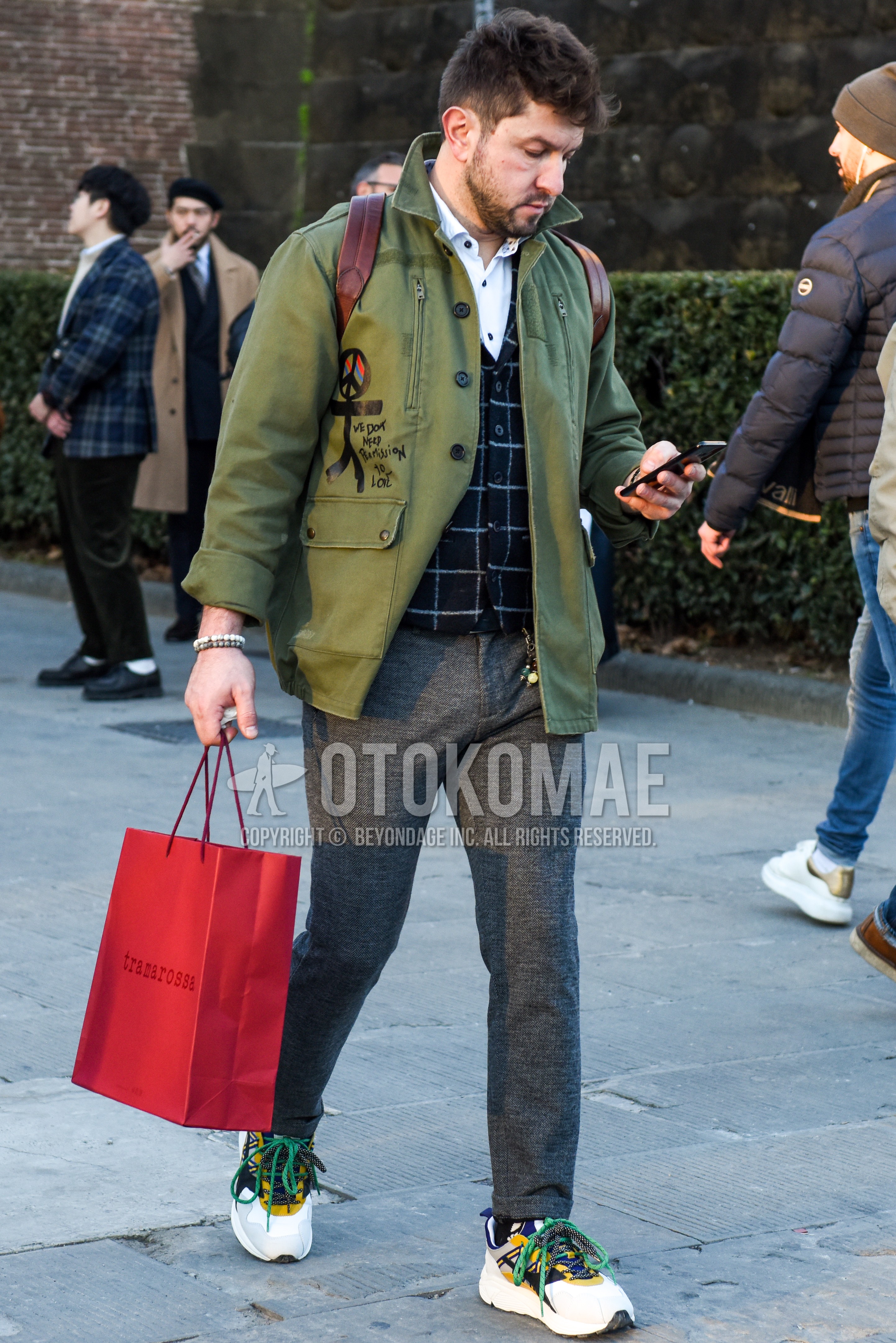Men's autumn winter outfit with olive green graphic field jacket/hunting jacket, navy check gilet, white plain shirt, gray plain slacks, white low-cut sneakers.