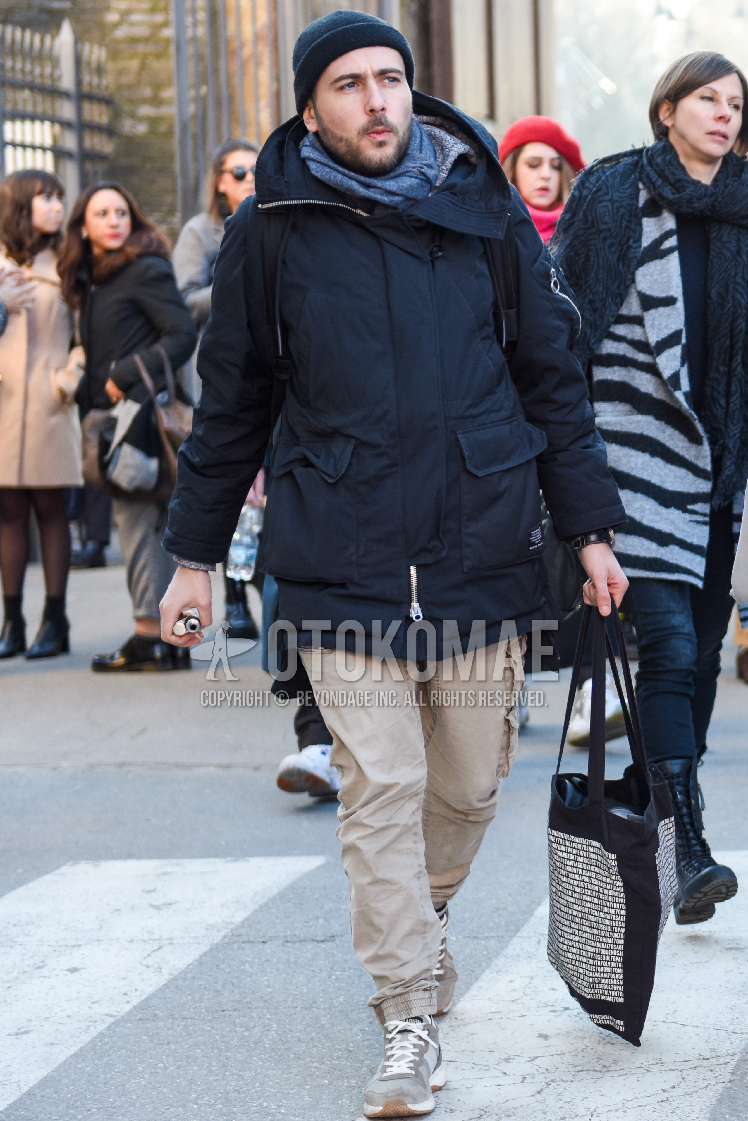 Men's autumn winter outfit with black plain knit cap, gray plain scarf, black plain hooded coat, beige plain chinos, beige plain cargo pants, gray low-cut sneakers, black graphic tote bag.