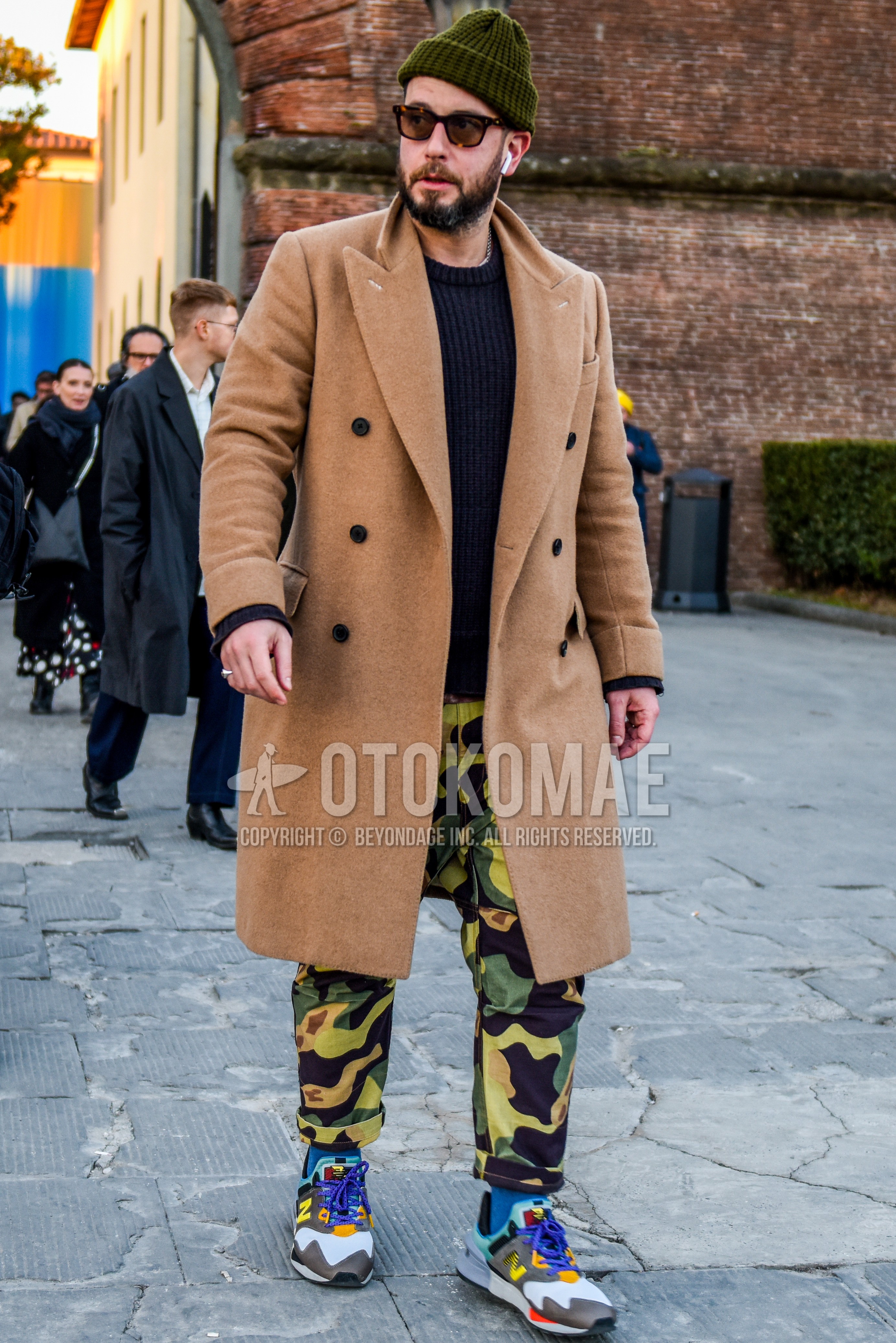 Men's autumn winter outfit with olive green plain knit cap, brown tortoiseshell sunglasses, beige plain chester coat, black plain sweater, green beige camouflage chinos, gray plain socks, gray low-cut sneakers.