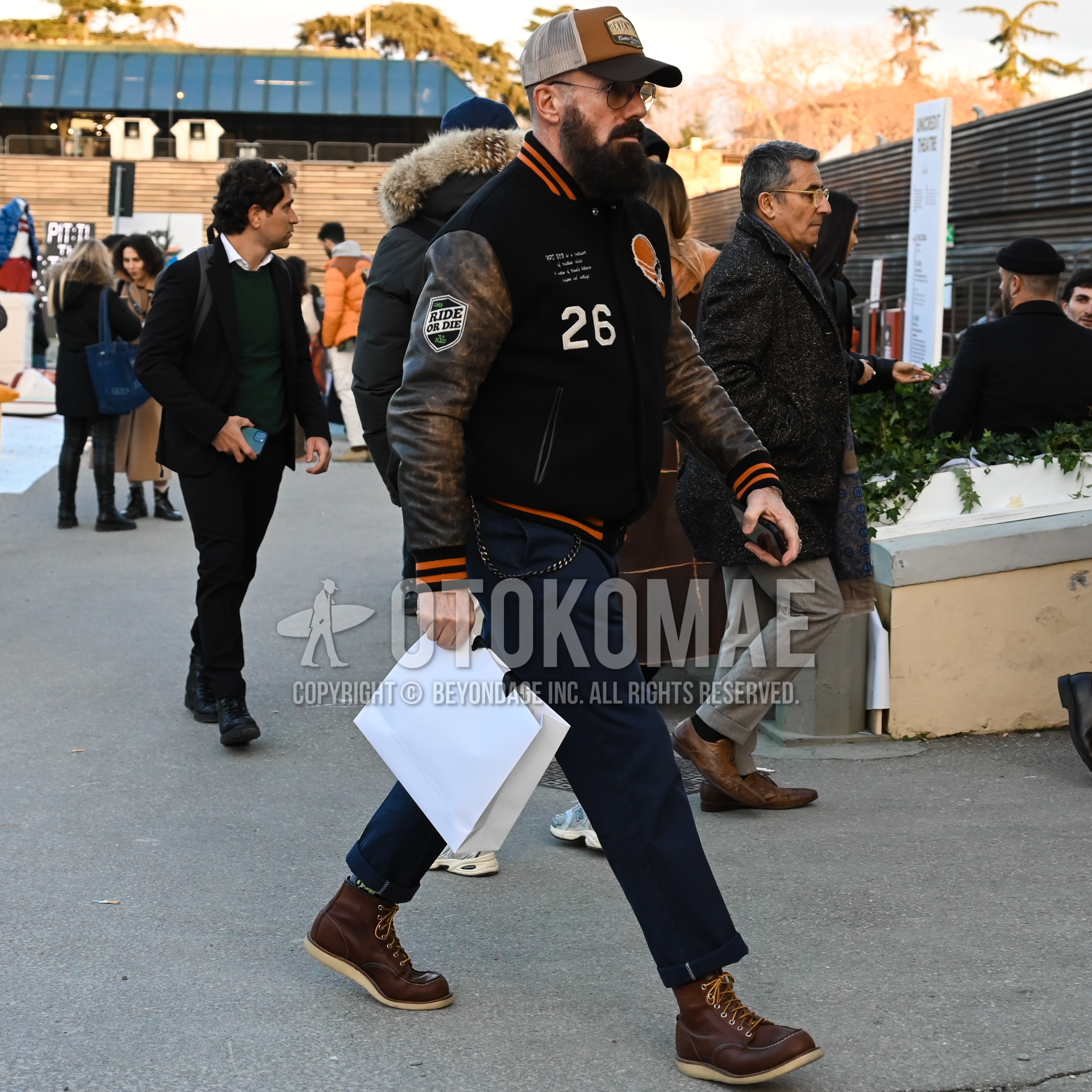 Men's autumn winter outfit with multi-color brown one point baseball cap, silver plain glasses, multi-color outerwear stadium jacket, navy plain ankle pants, brown work boots.