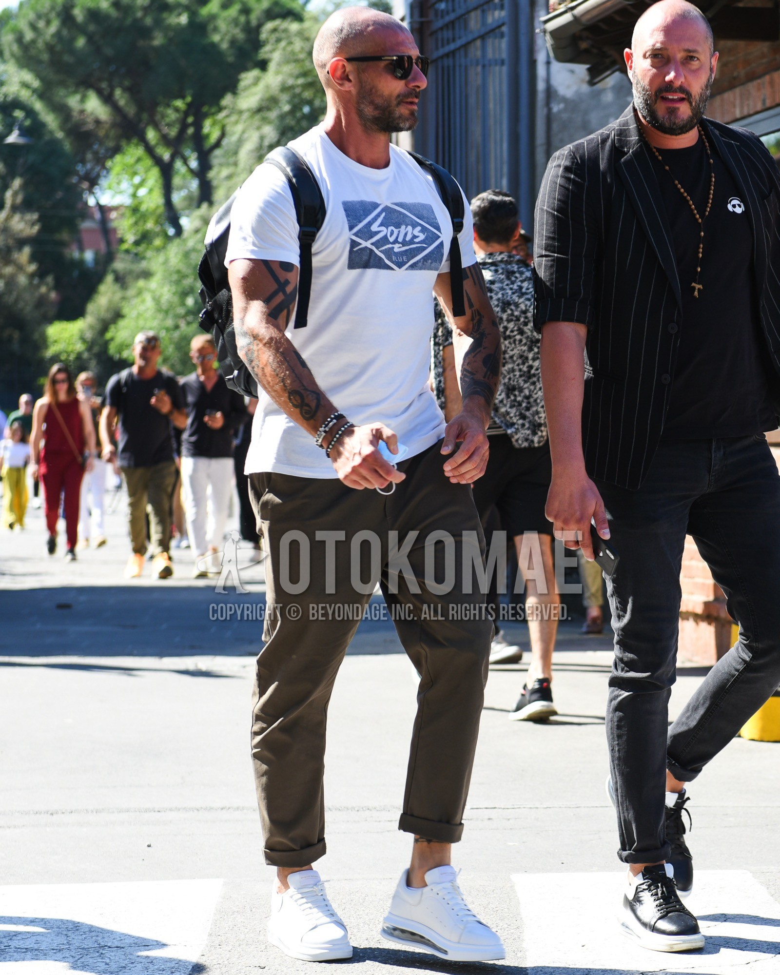 Men's spring summer outfit with black plain sunglasses, white graphic t-shirt, brown plain chinos, white low-cut sneakers, black plain backpack.