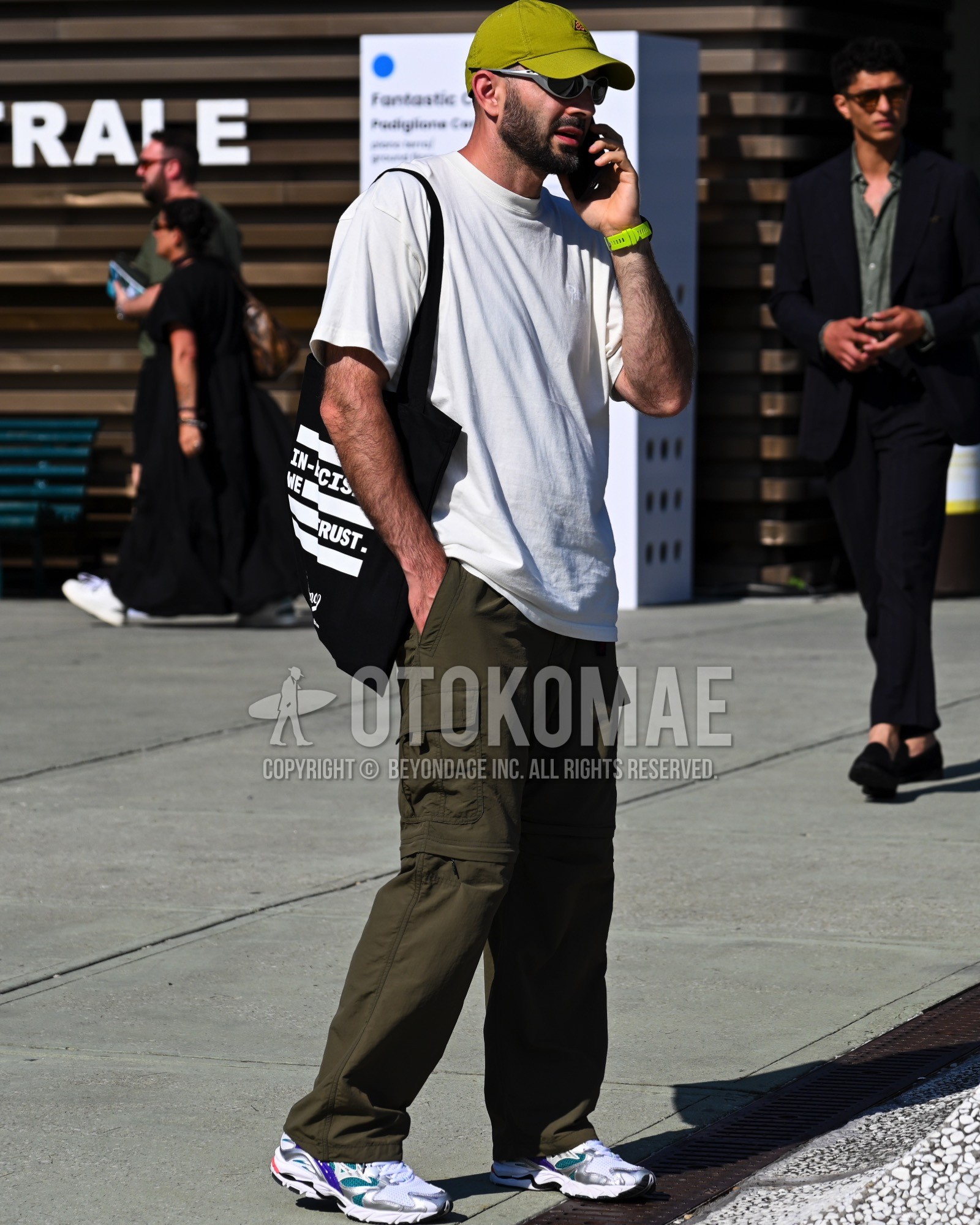 Men's spring summer outfit with green one point baseball cap, white plain t-shirt, olive green plain cargo pants, white low-cut sneakers, white graphic tote bag.